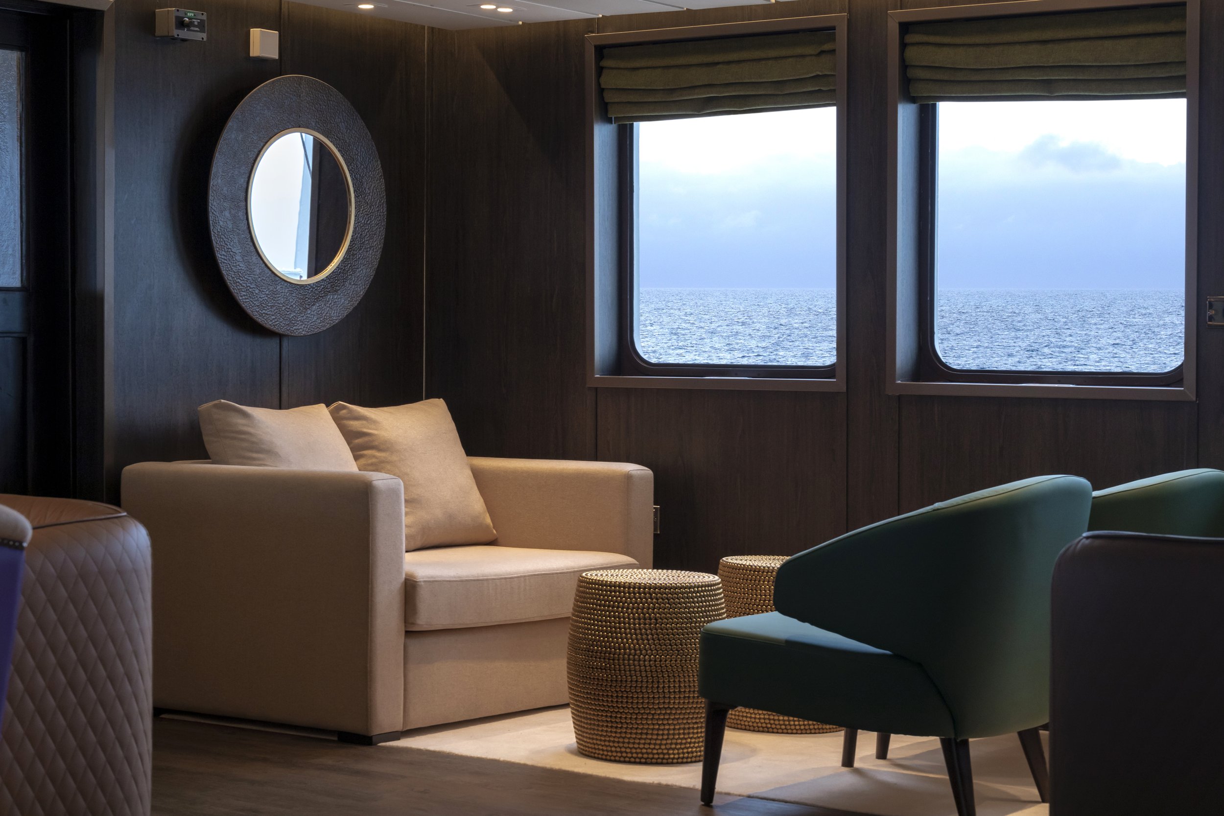 Luxurious room with dark wood paneled walls, two large windows overlooking the ocean, beige sofa with pillows, green and black chairs, and textured ottomans.