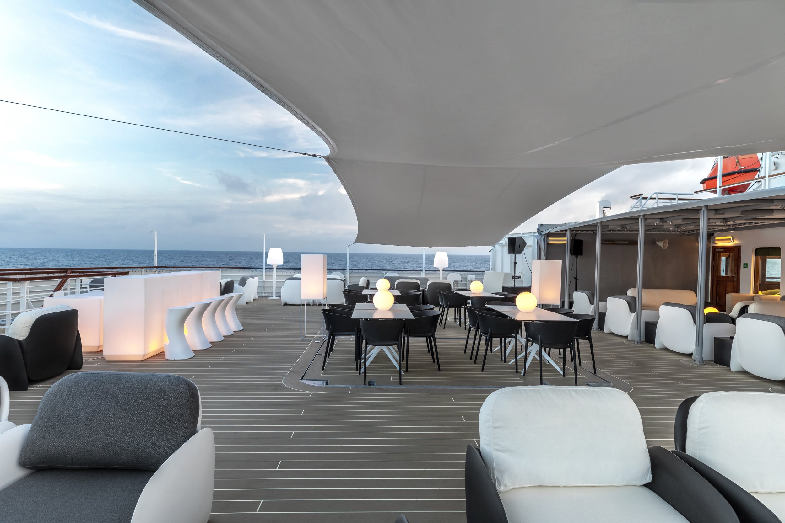 Empty outdoor lounge area on a cruise ship with black and white chairs, tables with glowing spherical lamps, and a view of the ocean in the background under a partially covered deck.