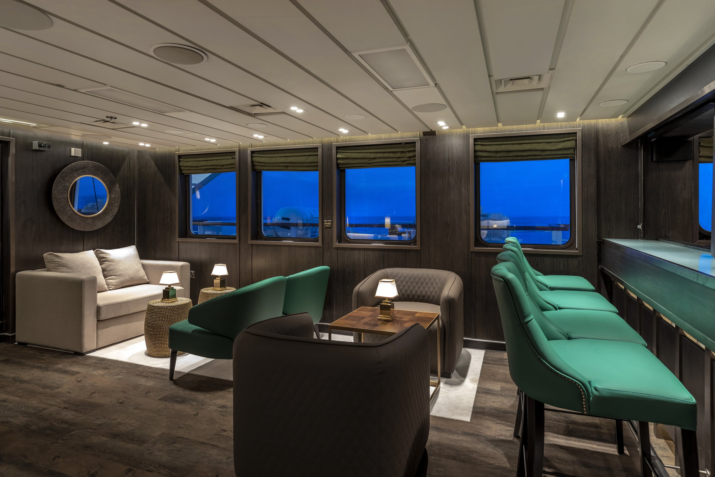 Elegant lounge area with a beige sofa, green chairs, and bar stools along a counter, illuminated by small table lamps and ceiling lights, with a dark wood wall and large windows showing an evening view outside.