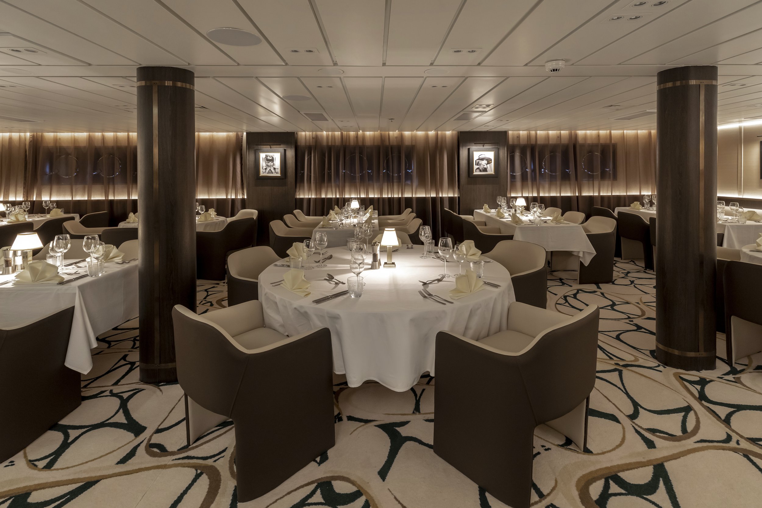 Elegant dining room with round tables covered in white tablecloths, set with glassware, silverware, and napkins, surrounded by cushioned chairs, featuring warm lighting and dark wood accents.