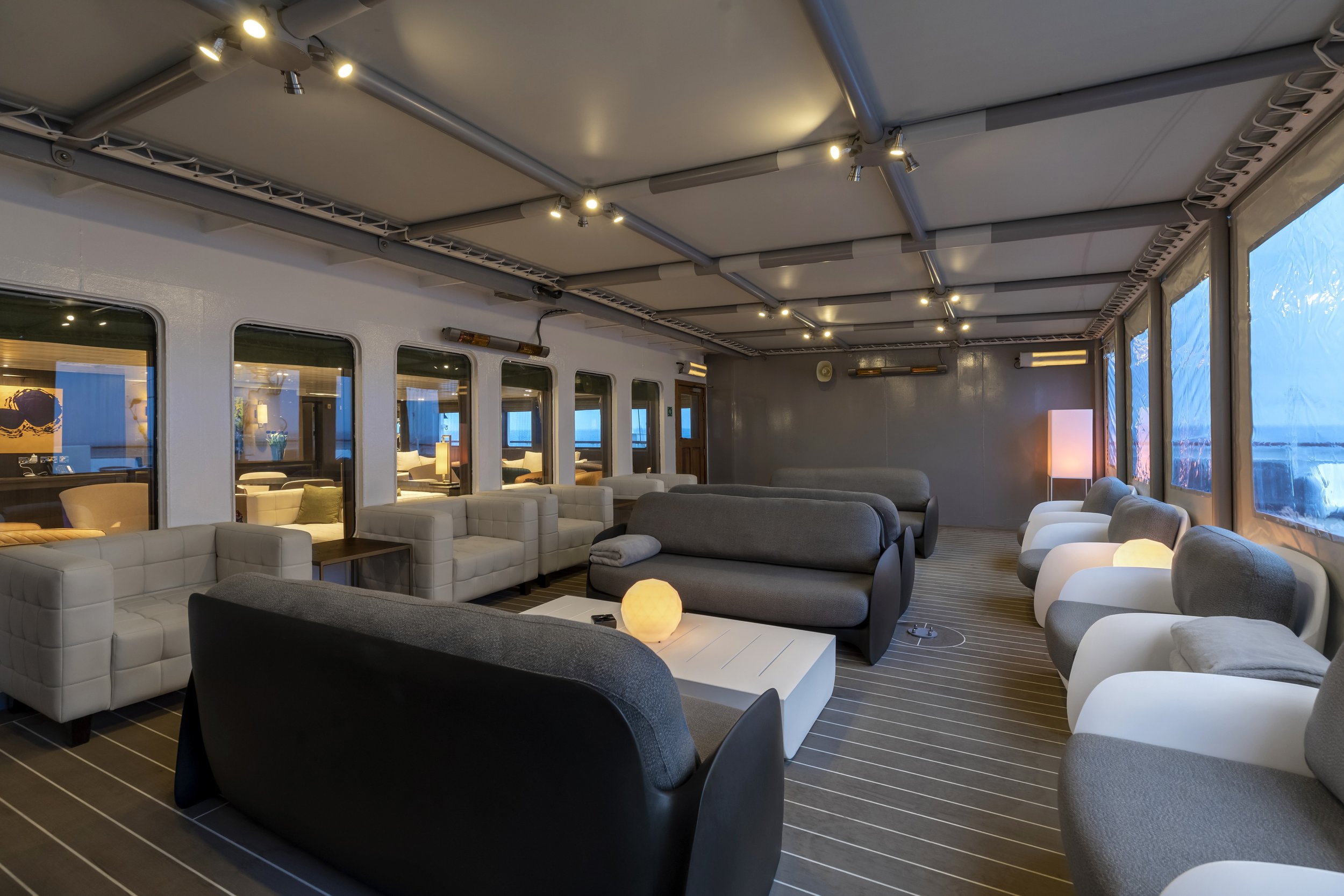 Interior of a lounge area with various modern chairs and sofas, low tables, and soft lighting. Large windows with blue-tinted views, and a decorated ceiling with track lighting.