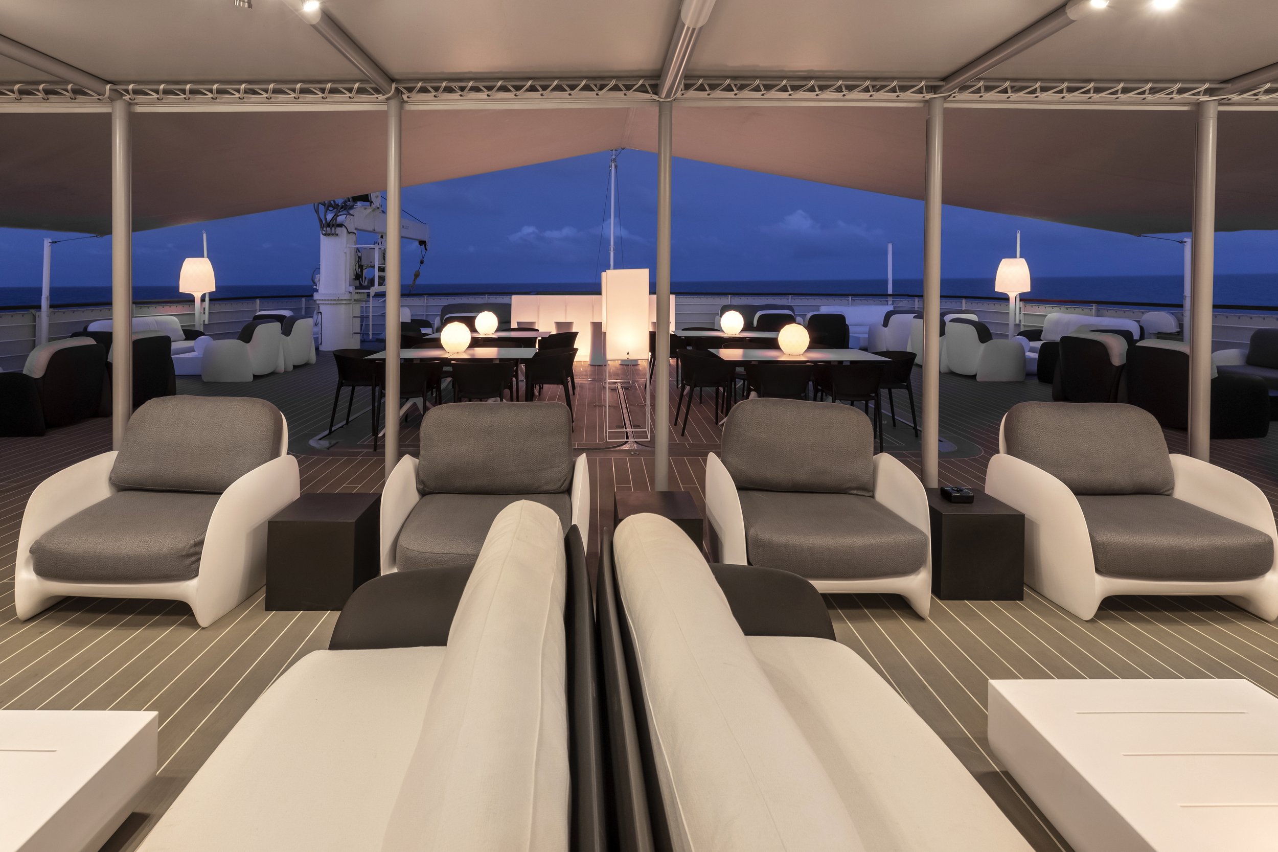 Luxury outdoor lounge area on a cruise ship rooftop deck at night, with modern white and gray chairs, black tables, lamps, and a view of the ocean in the background.