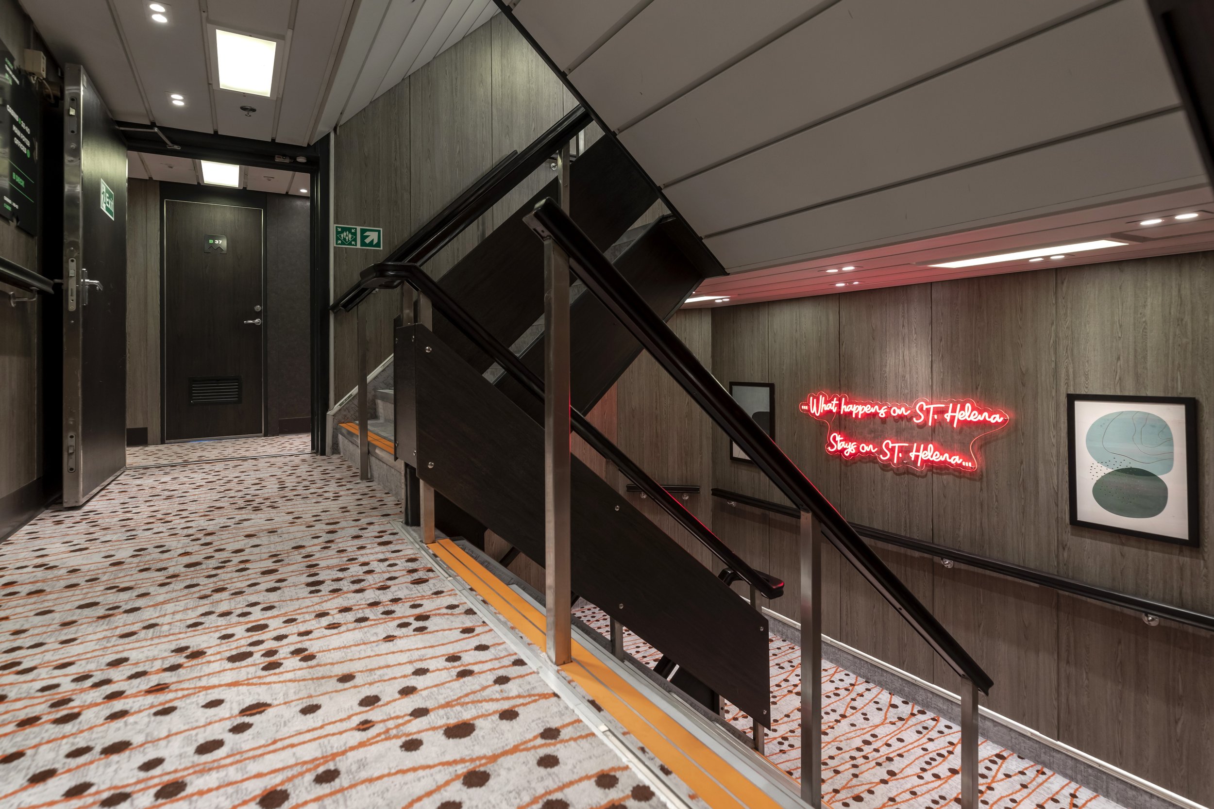 Interior view of a building corridor with a staircase, wooden walls, patterned carpet, and framed artwork. There is a neon sign on the wall that reads 'What happens on ST. Helena stays on ST. Helena'.