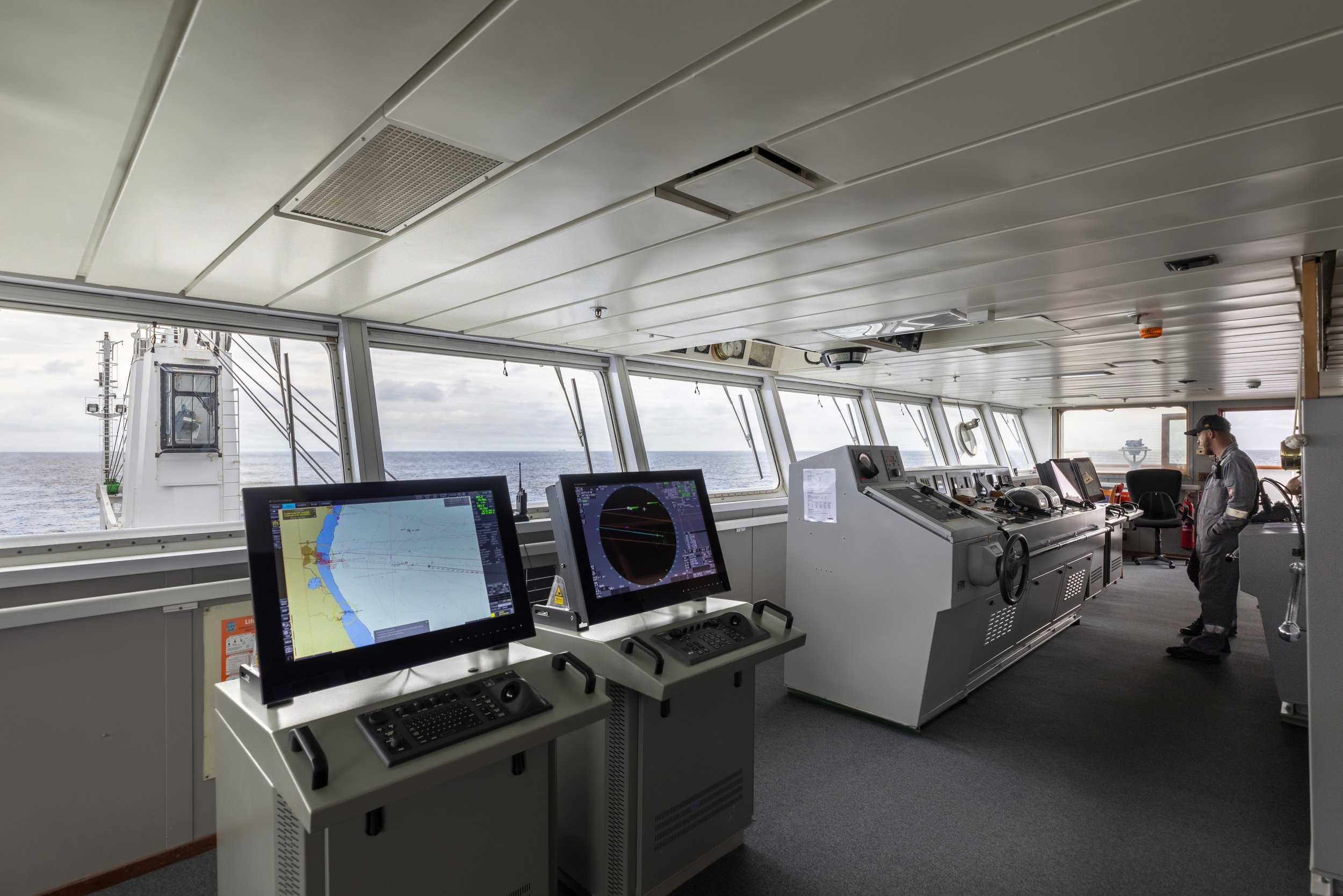Ship bridge with navigation and control screens, window views of the ocean, and crew member standing.