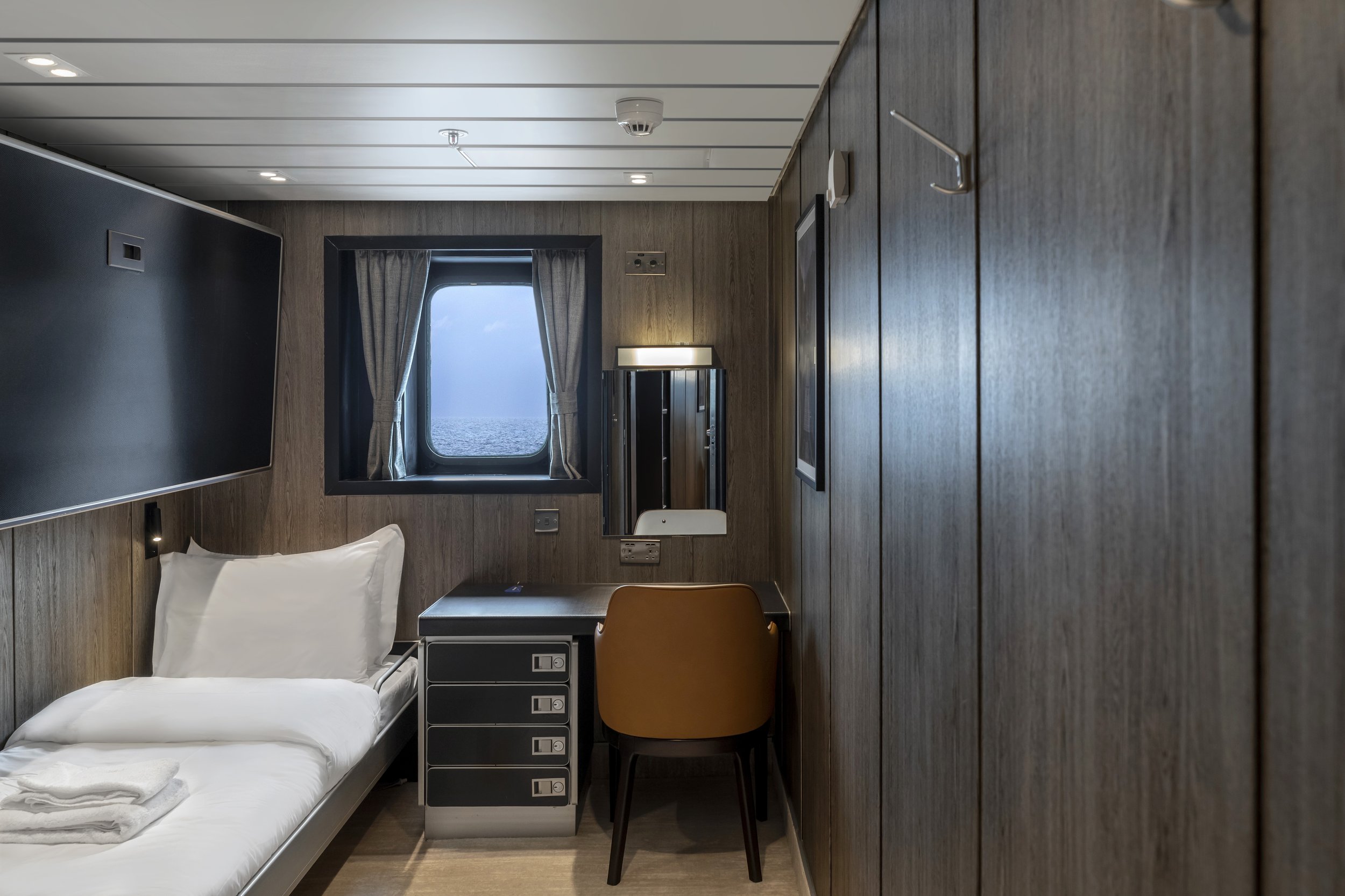 Small hotel room with a single bed, window with ocean view, black wall-mounted TV, desk with a chair, and wooden cabinets.