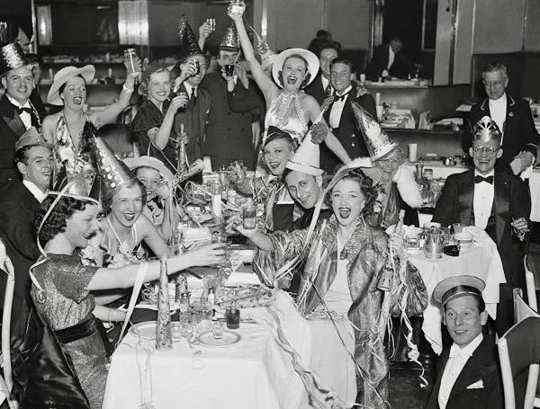 Group of people celebrating a party, wearing hats and party accessories, sitting around a table with drinks and confetti, in a banquet hall.