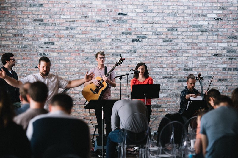 A band performing in front of an exposed brick wall with a man playing guitar, a woman singing into a microphone, and another person playing a cello. People in front appear to be worshiping or listening.