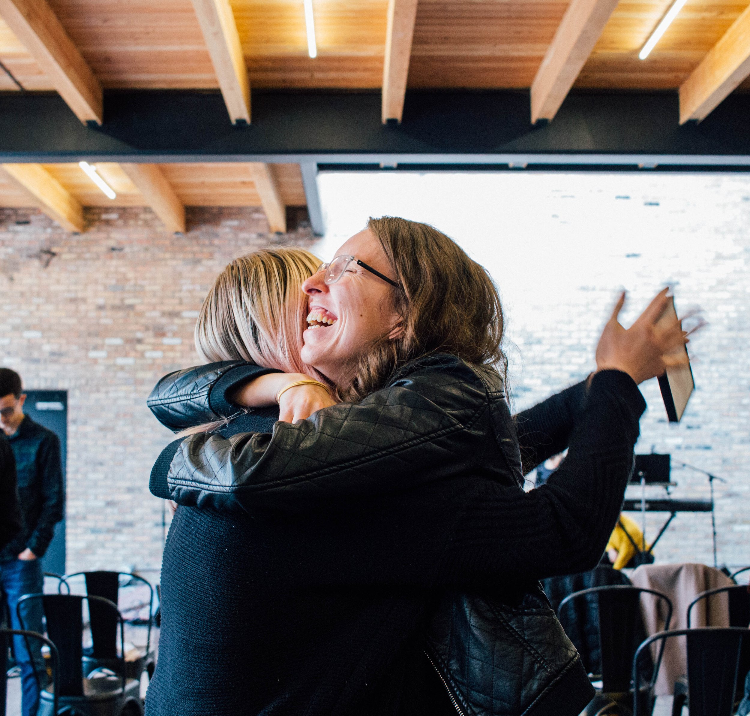 Two women hugging and smiling in a room with wooden beams and exposed brick walls.
