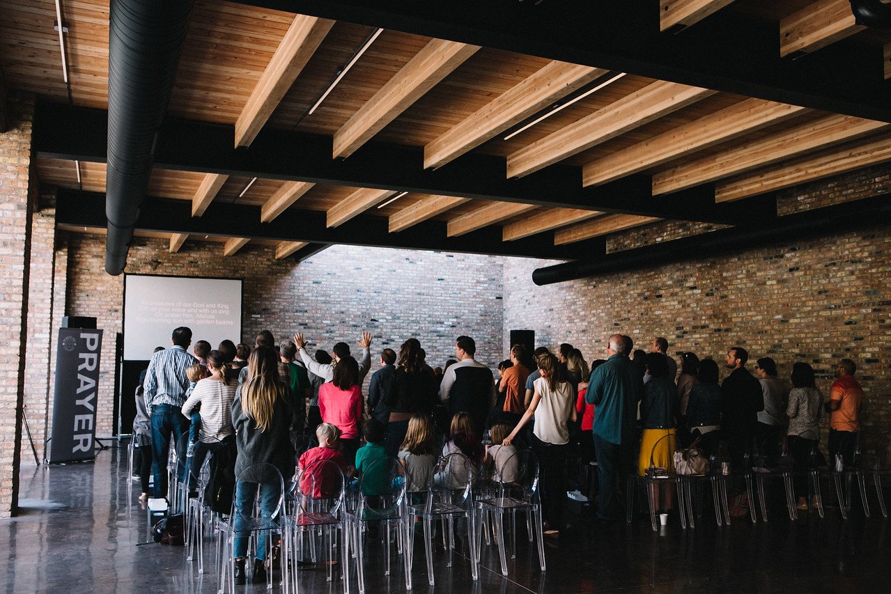 Group of people gathered for a religious service in a brick-walled venue, facing a screen with song lyrics. "Prayer" banner visible.