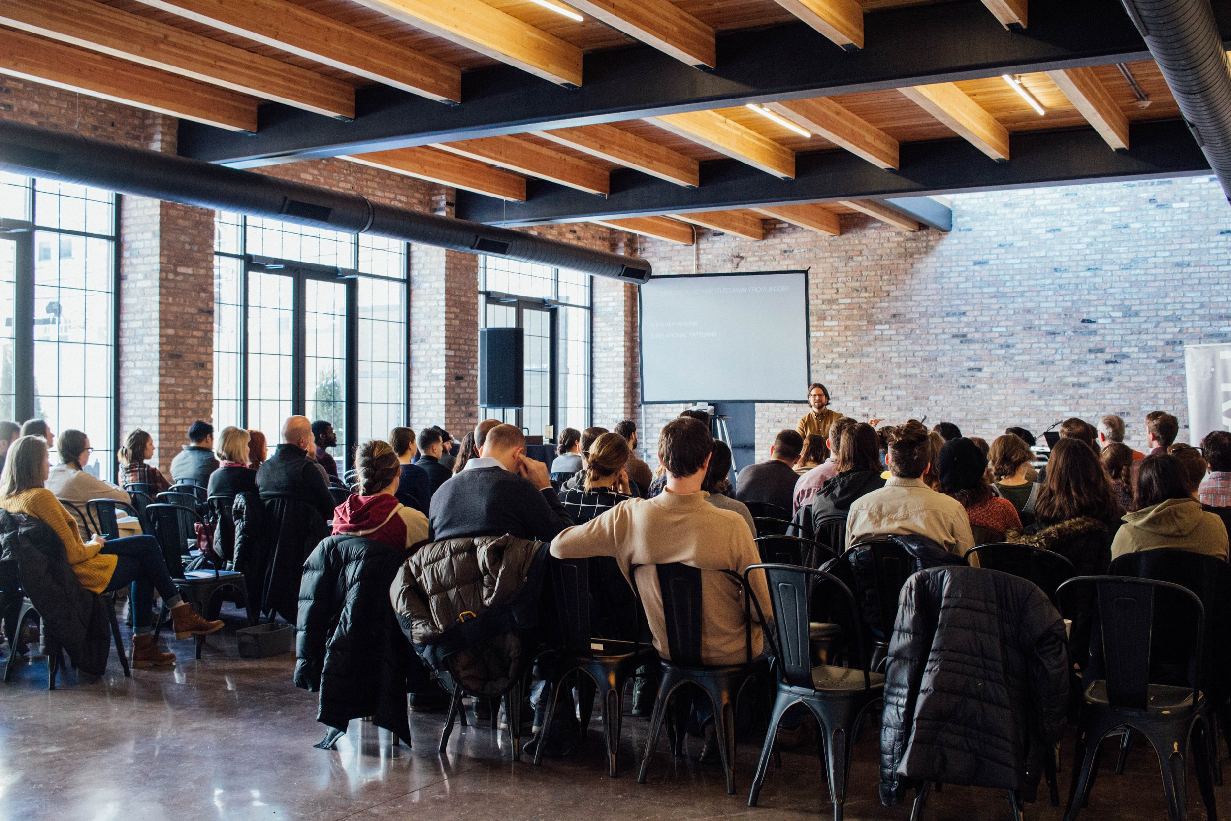 A group of people attending a seminar in a room with exposed beams and brick walls. The audience is seated, facing a presenter in front of a projector screen.