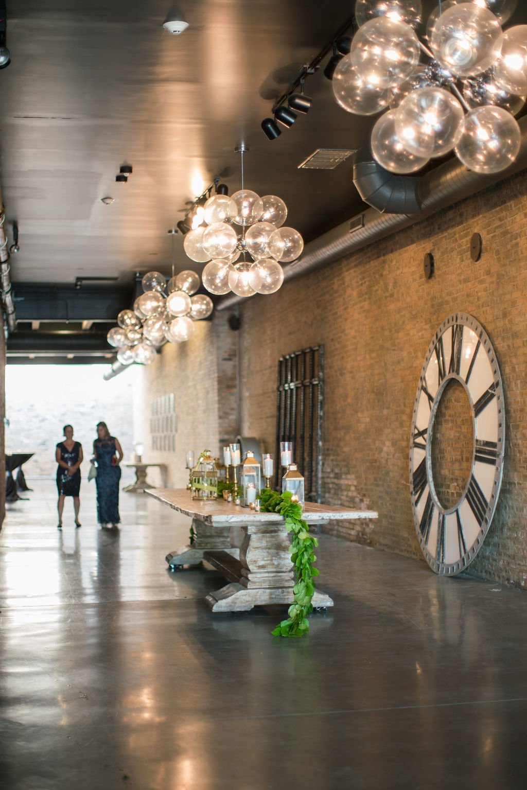 Interior of a modern hallway with brick walls, large clock face, and decorative lighting fixtures. A long table with candles and greenery in the foreground. Two people in formal attire stand in the background.