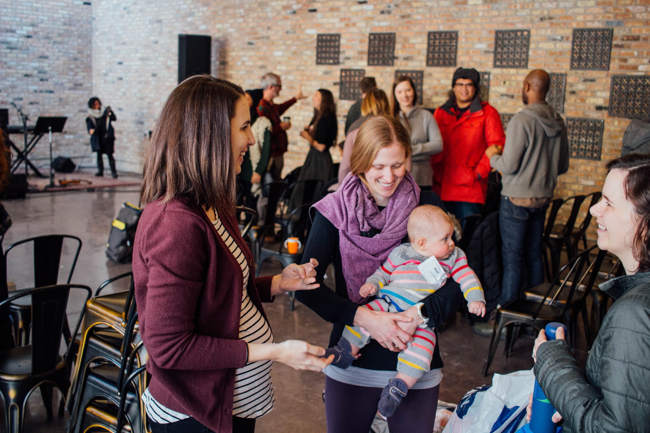 Group of people in a casual indoor gathering, two women smiling at each other, one holding a baby in colorful clothes, with others interacting in the background, brick wall setting.