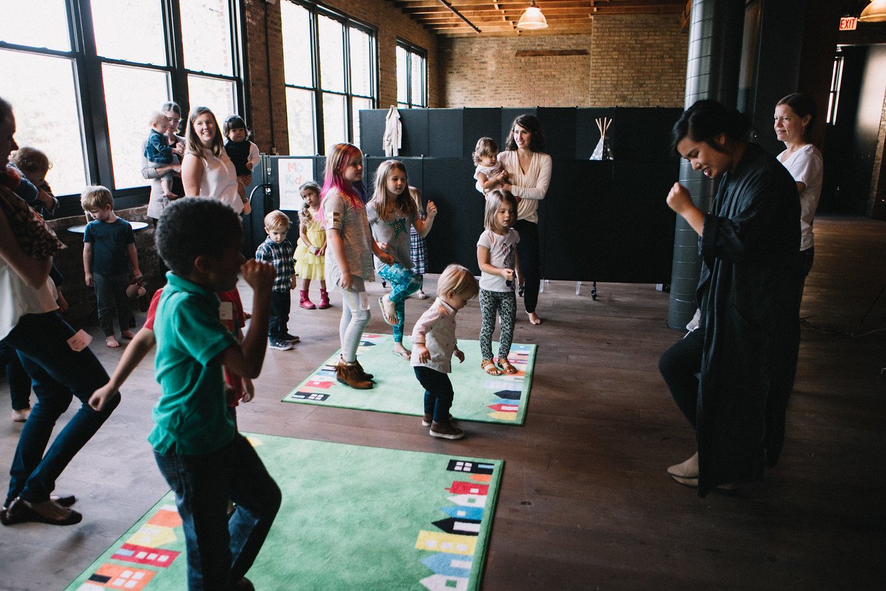 Children and adults participating in an indoor dance activity, with some children standing on colorful mats, in a room with large windows and wooden floors.