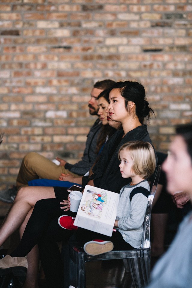 Group of people sitting in rows listening attentively, with a child holding an open book.