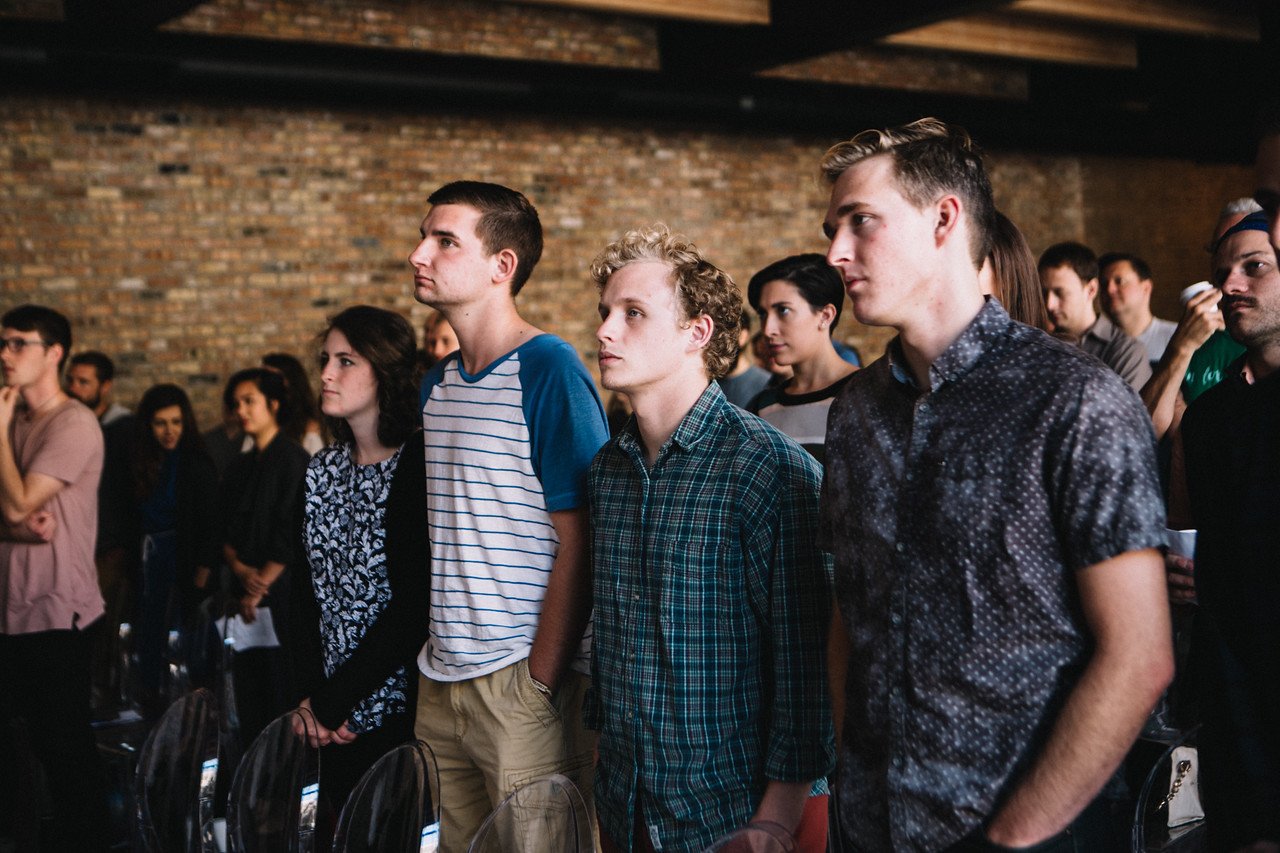 Group of people attentively listening indoors, standing in rows in a brick-walled room.