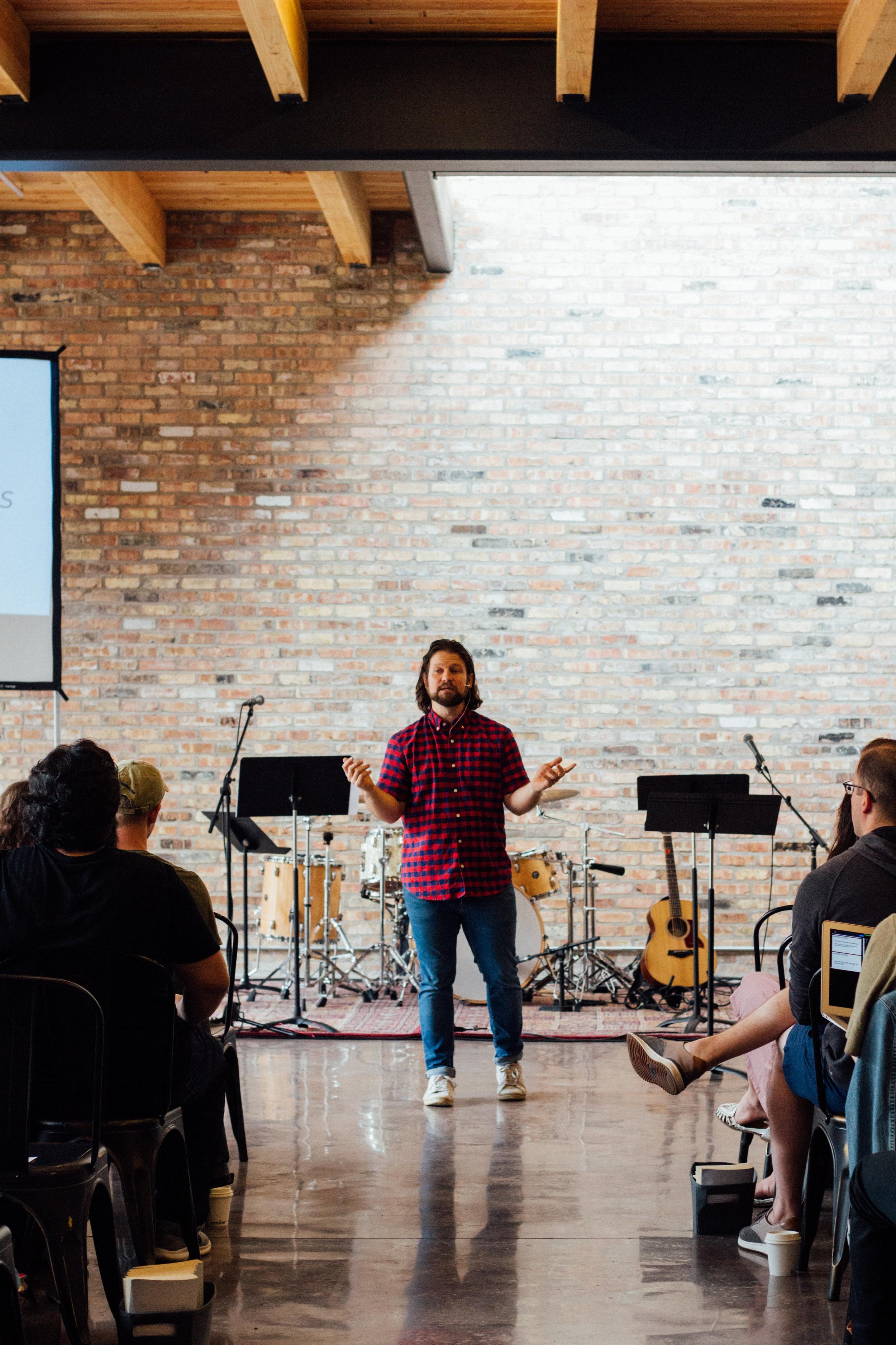 A person speaking to an audience in a casual setting, with musical instruments in the background, including a drum set and a guitar.