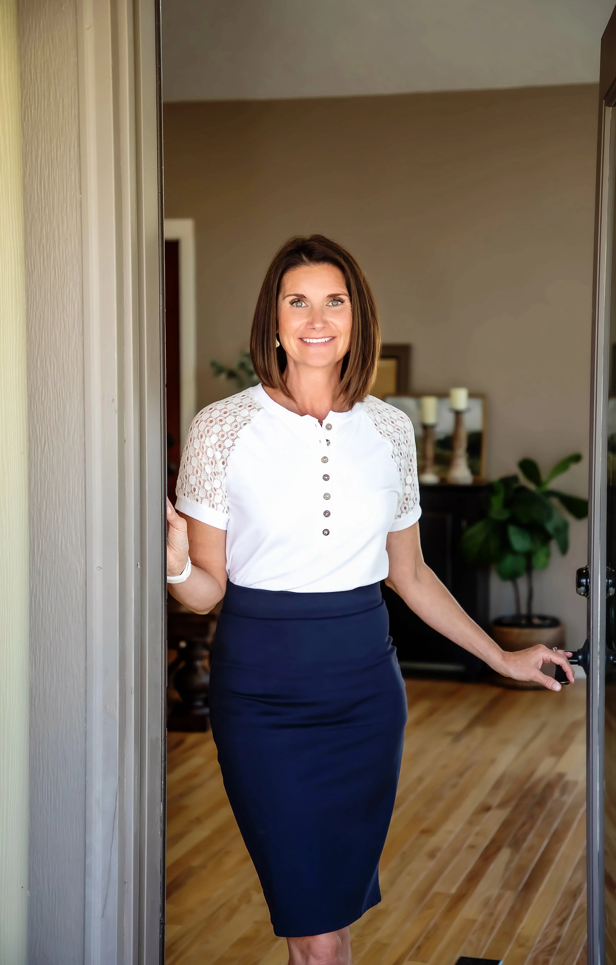 A woman standing at the doorway, smiling, wearing a white top with lace sleeves and a dark blue pencil skirt in a home interior with hardwood flooring and plants in the background.