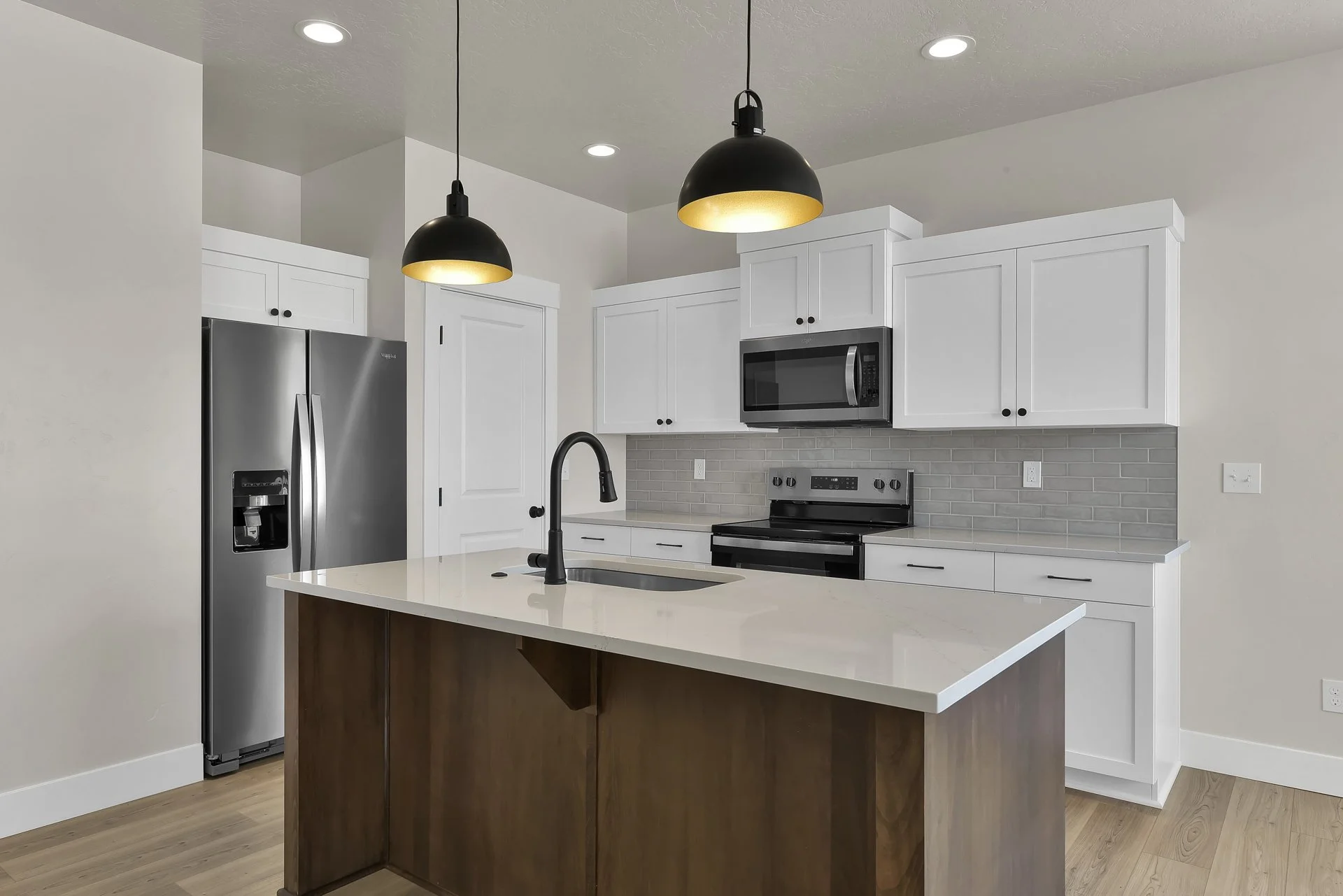 Modern kitchen with white cabinets, stainless steel refrigerator, microwave, electric stove, gray tile backsplash, island with white countertop, black faucet, and black pendant lighting.