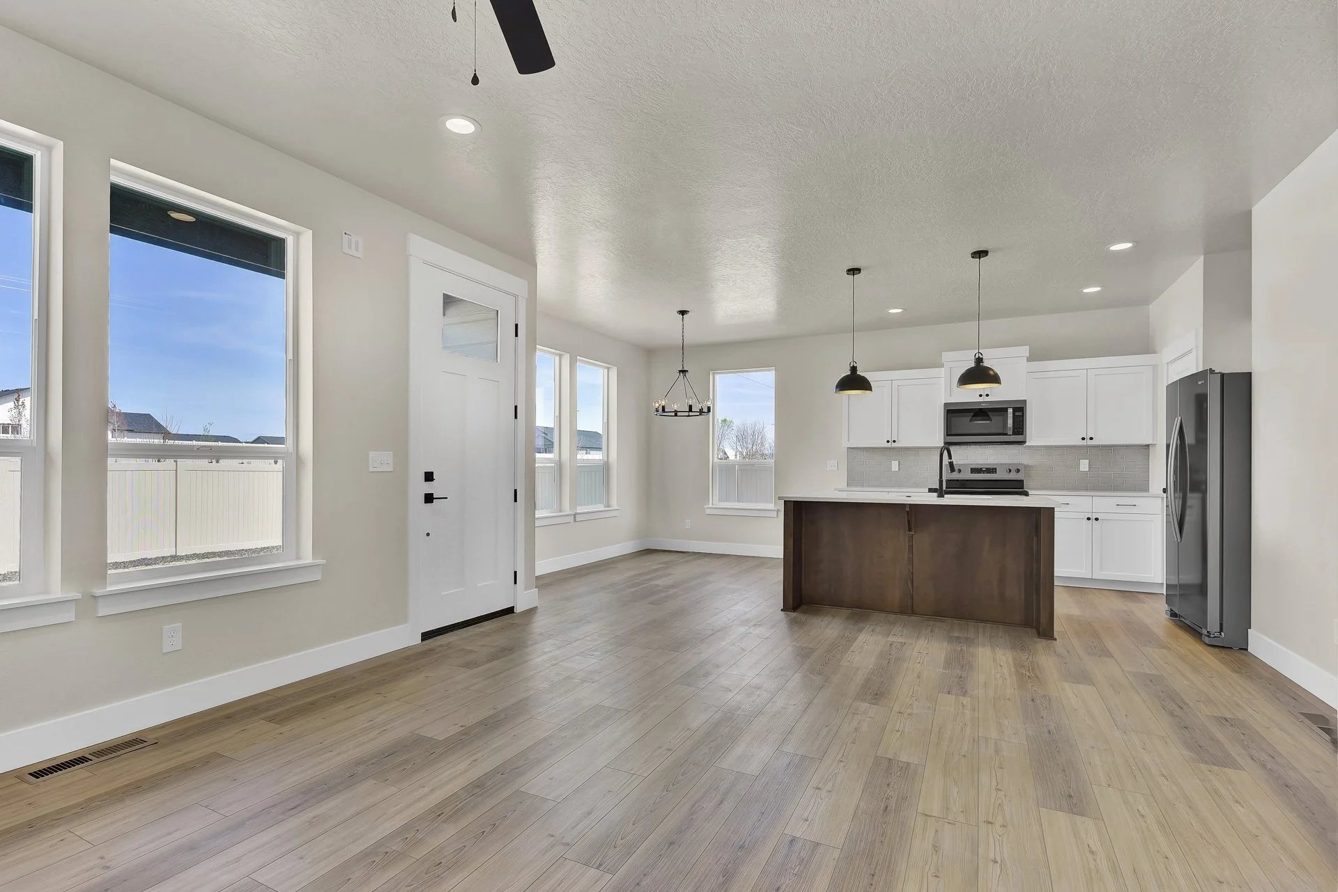 Empty modern kitchen and dining area with hardwood floors, white cabinets, stainless steel appliances, black pendant lights, and large windows letting in natural light.