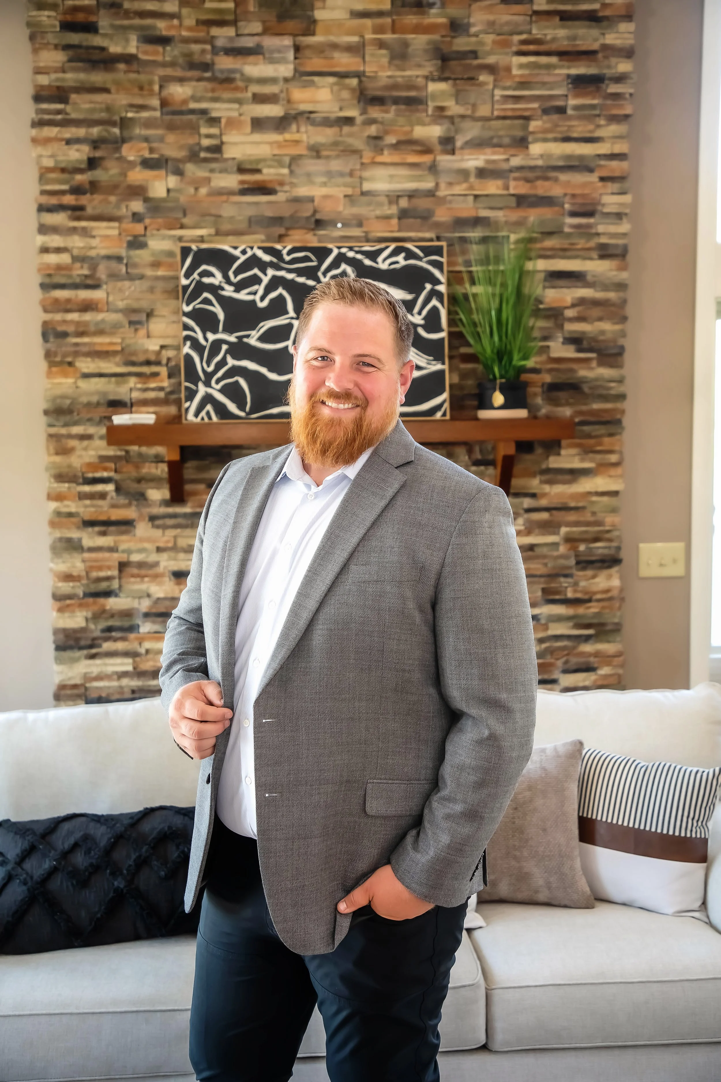 A man with a beard in a gray blazer and white shirt smiling in a living room with a stone accent wall, a sofa with pillows, and a bookshelf