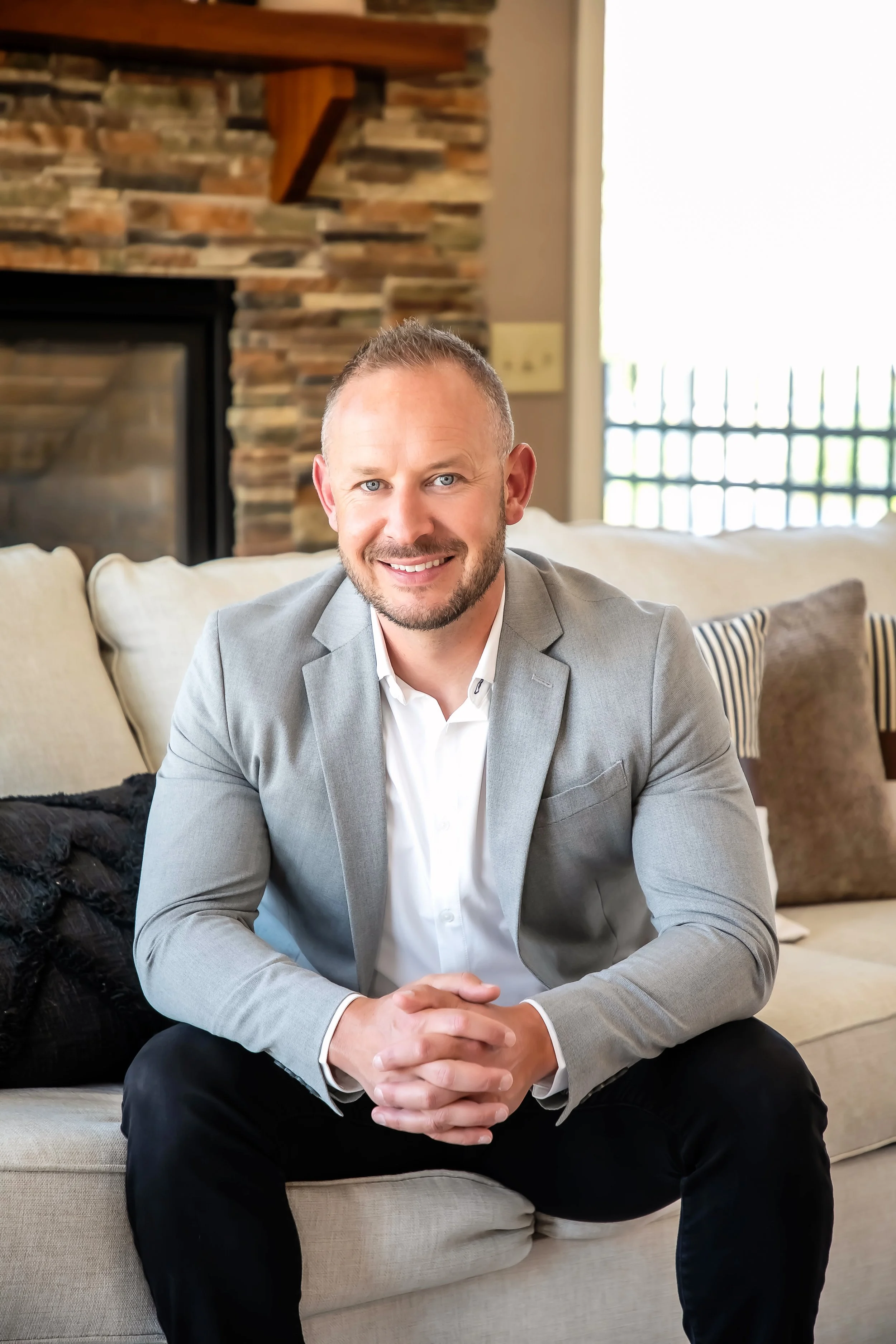 A smiling man in a gray blazer and white shirt sitting on a beige sofa in a living room with a stone fireplace and a window in the background.
