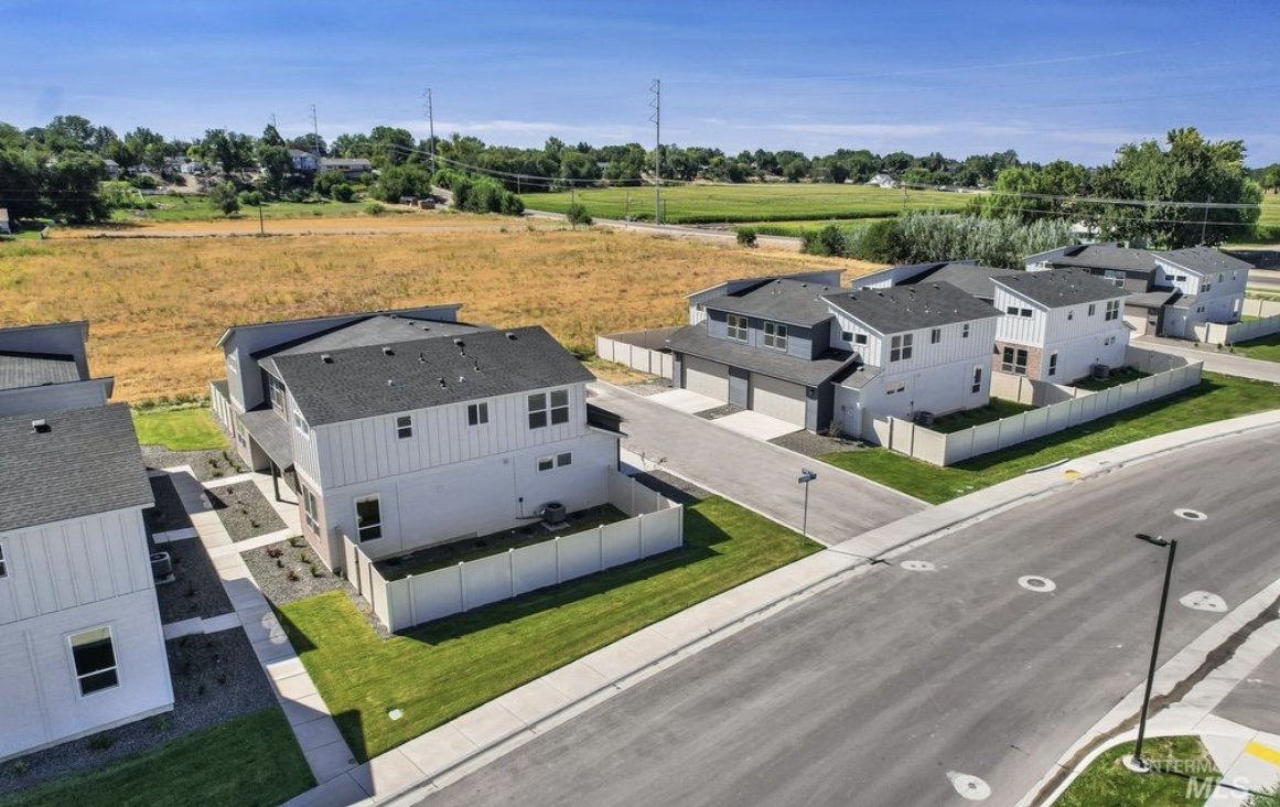 Aerial view of newly constructed modern houses with white exteriors, dark roofs, and fenced backyards in a suburban neighborhood, with empty roads and streetlights, and fields and trees in the background.