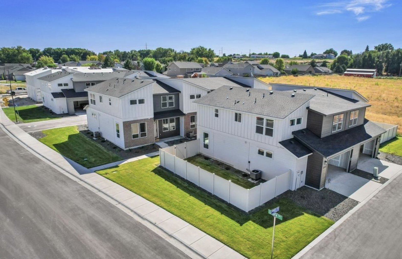 Aerial view of new modern residential houses with white and gray exteriors, manicured lawns, driveways, and a white fence, in a suburban neighborhood under a bright blue sky.