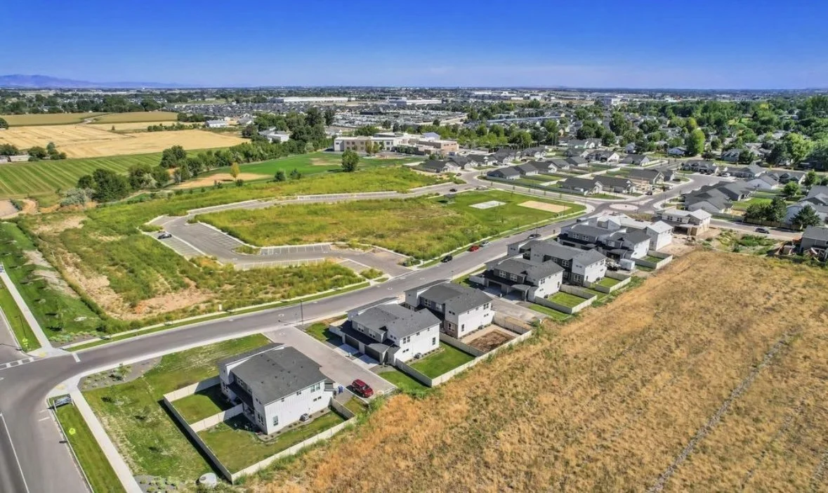 Aerial view of a suburban neighborhood with newly built houses, green lawns, and open land under development, surrounding agricultural fields and distant cityscape under a clear blue sky.