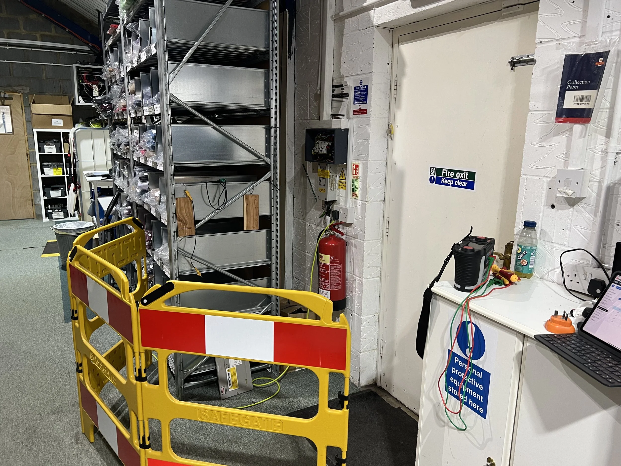 A storage room with metal shelving units filled with boxes and equipment. A yellow and red safety barrier is in front. There is a fire extinguisher mounted on the wall near a closed white door that has a sign indicating it is a fire exit with a keep clear notice. On the right, a white counter holds some tools, a bottle of water, a laptop, and electrical cords.