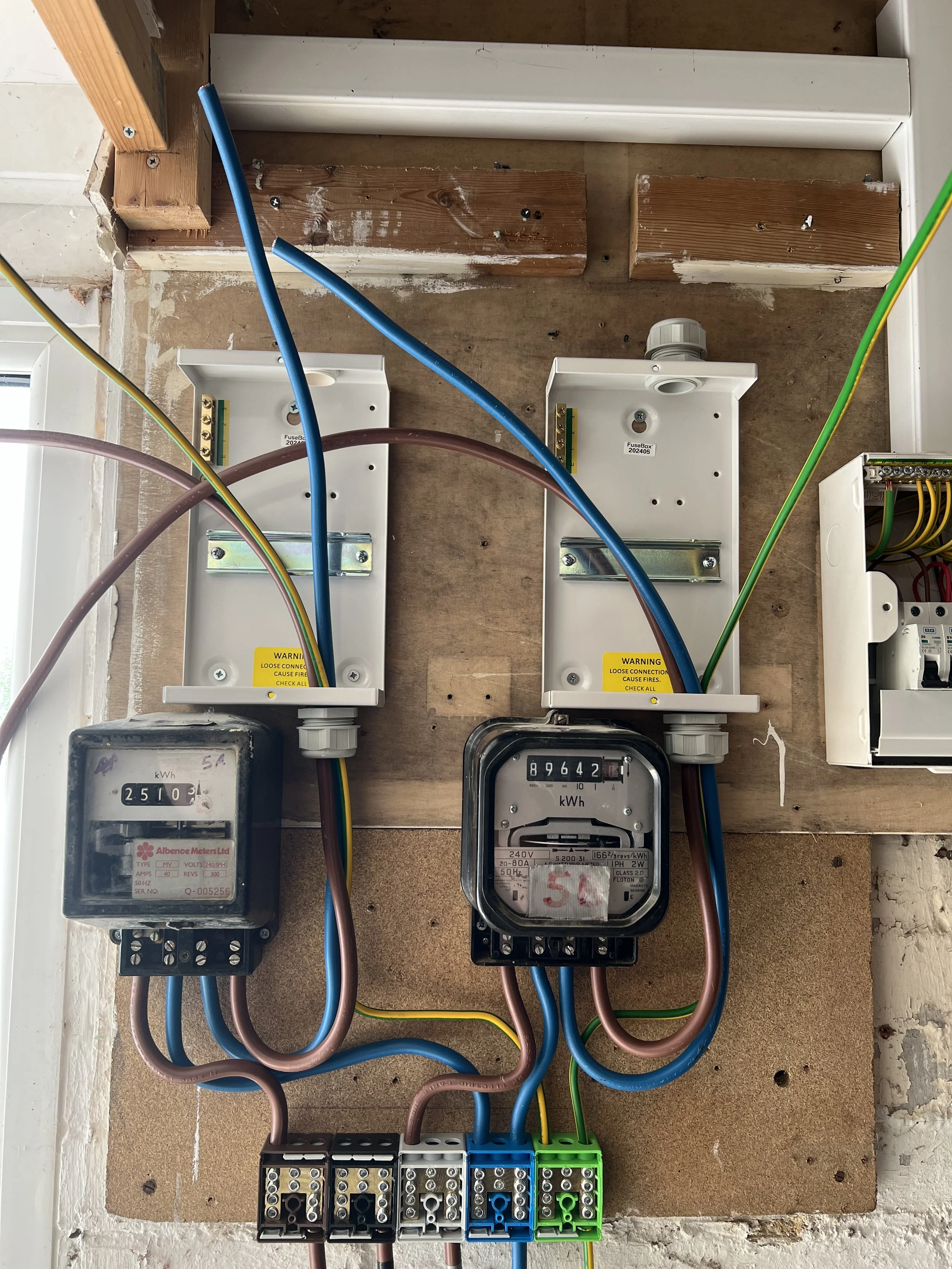Electrical meters and wiring mounted on a wooden panel with multiple colored wires connected to different terminals and boxes.