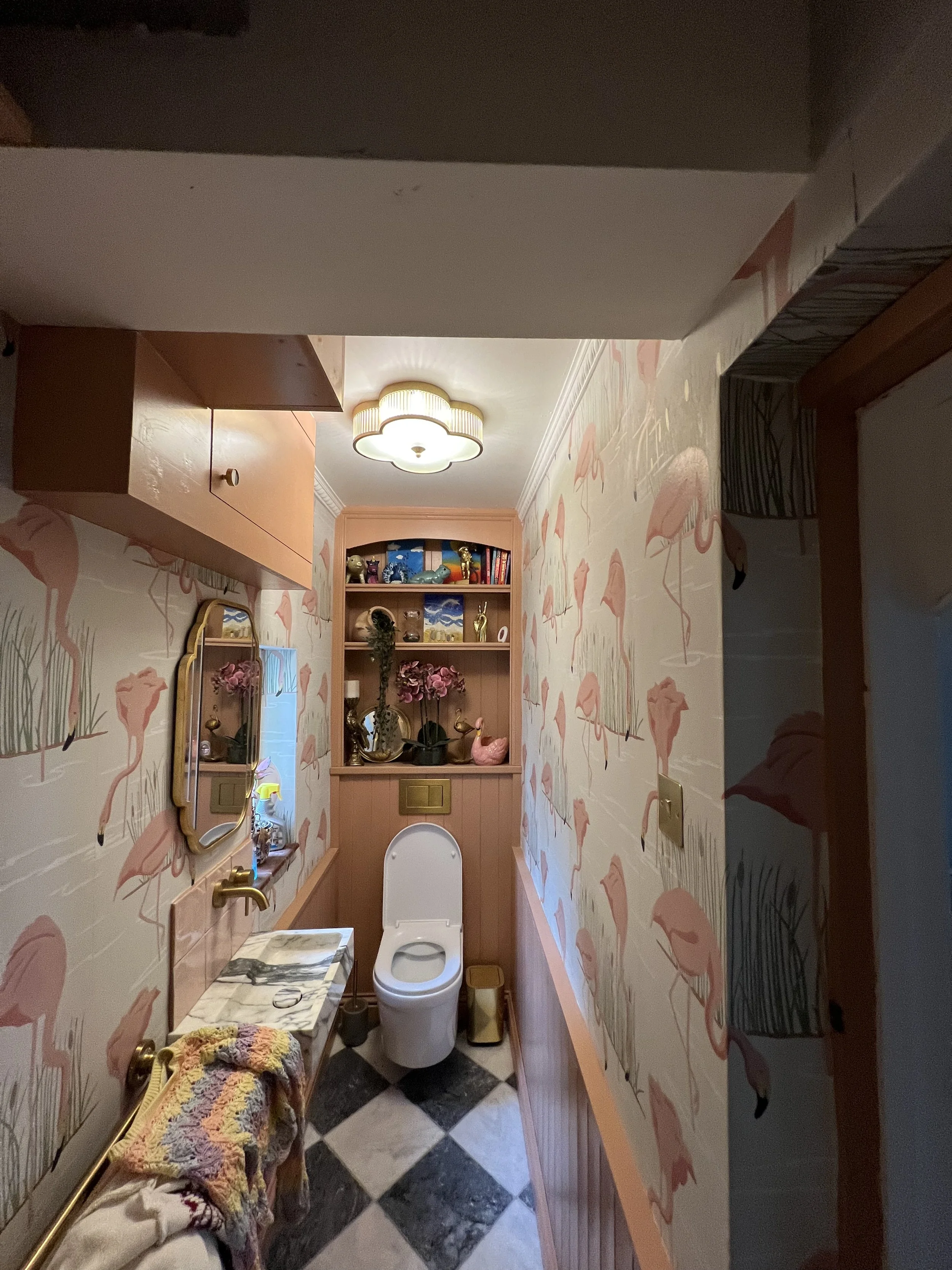 A small bathroom decorated with flamingo-themed wallpaper, pink wooden paneling, and flamingo figurines on a shelf. It has a white toilet, a marble-topped vanity, a mirror, and a beige ceiling light fixture. The floor features black and white diamond tiles.