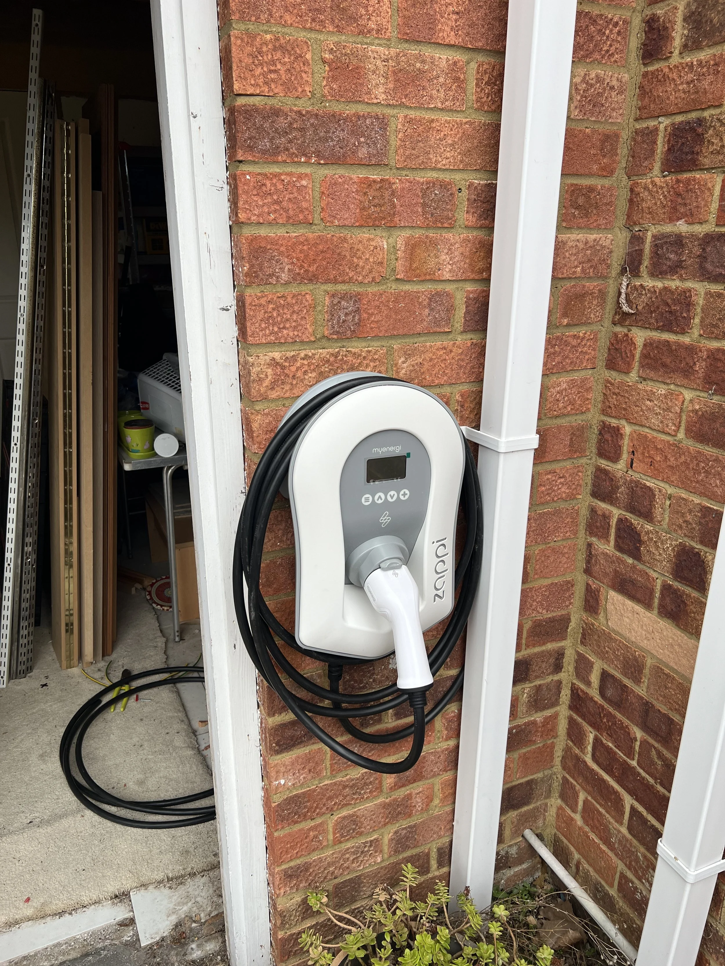 An electric vehicle charger installed on a brick wall outside a building, with a coiled black cable hanging from it.