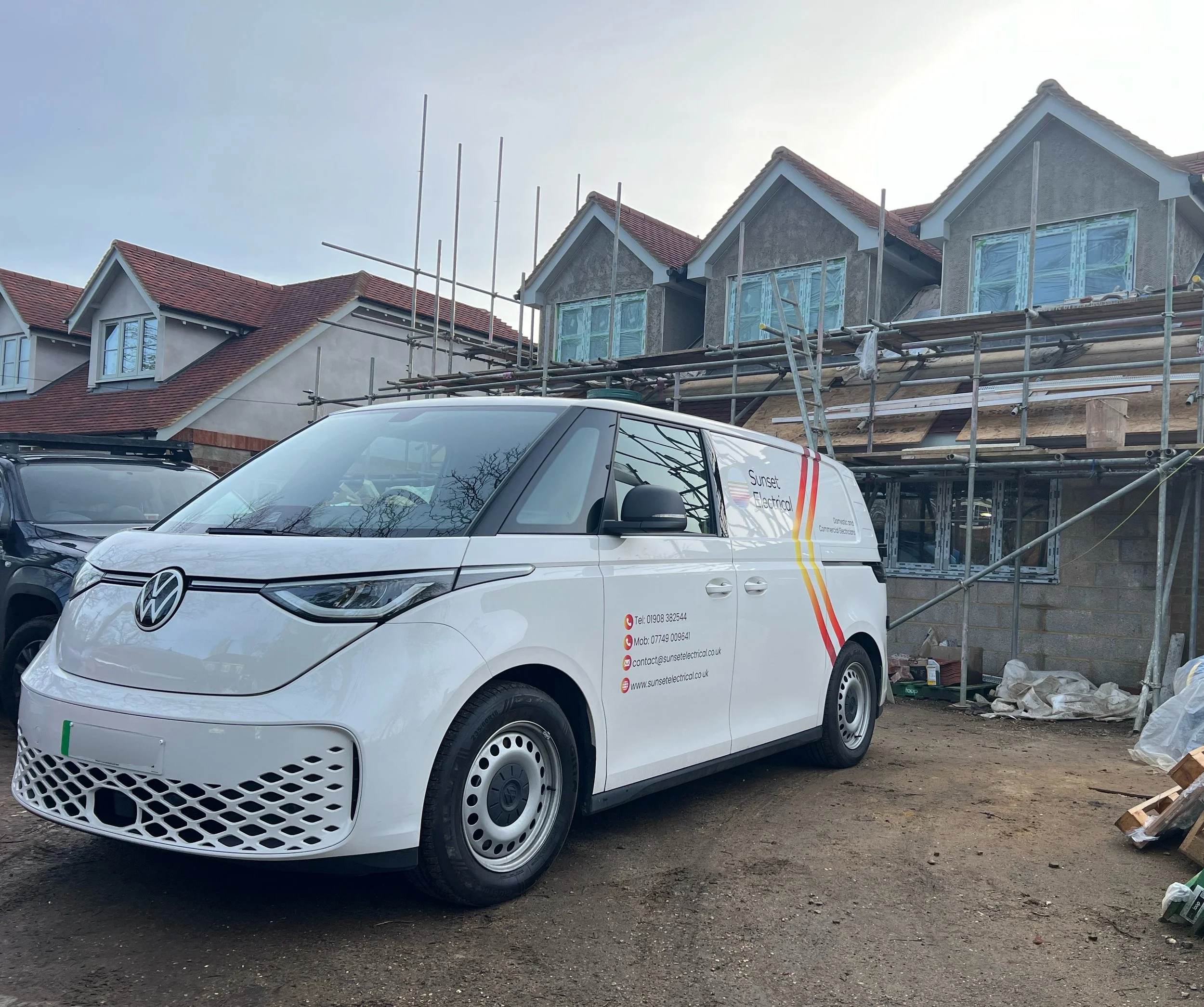 Construction site with scaffolding and partially built houses, white electrical work van parked in front, overcast sky.