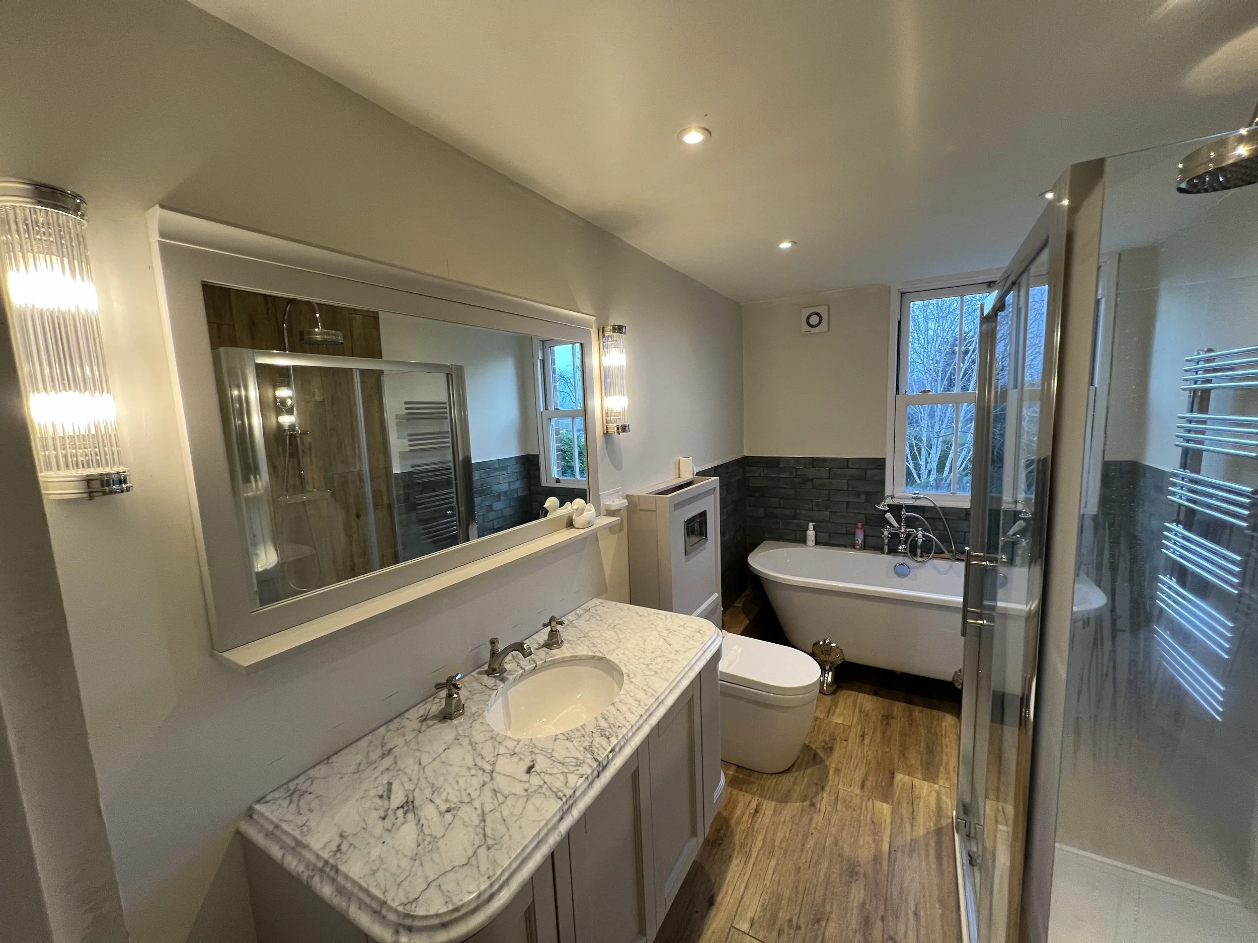 A modern bathroom with a marble sink vanity, a large mirror, a toilet, a bathtub under a window, and a shower stall with glass door. The room has wood flooring and decorative wall lighting.