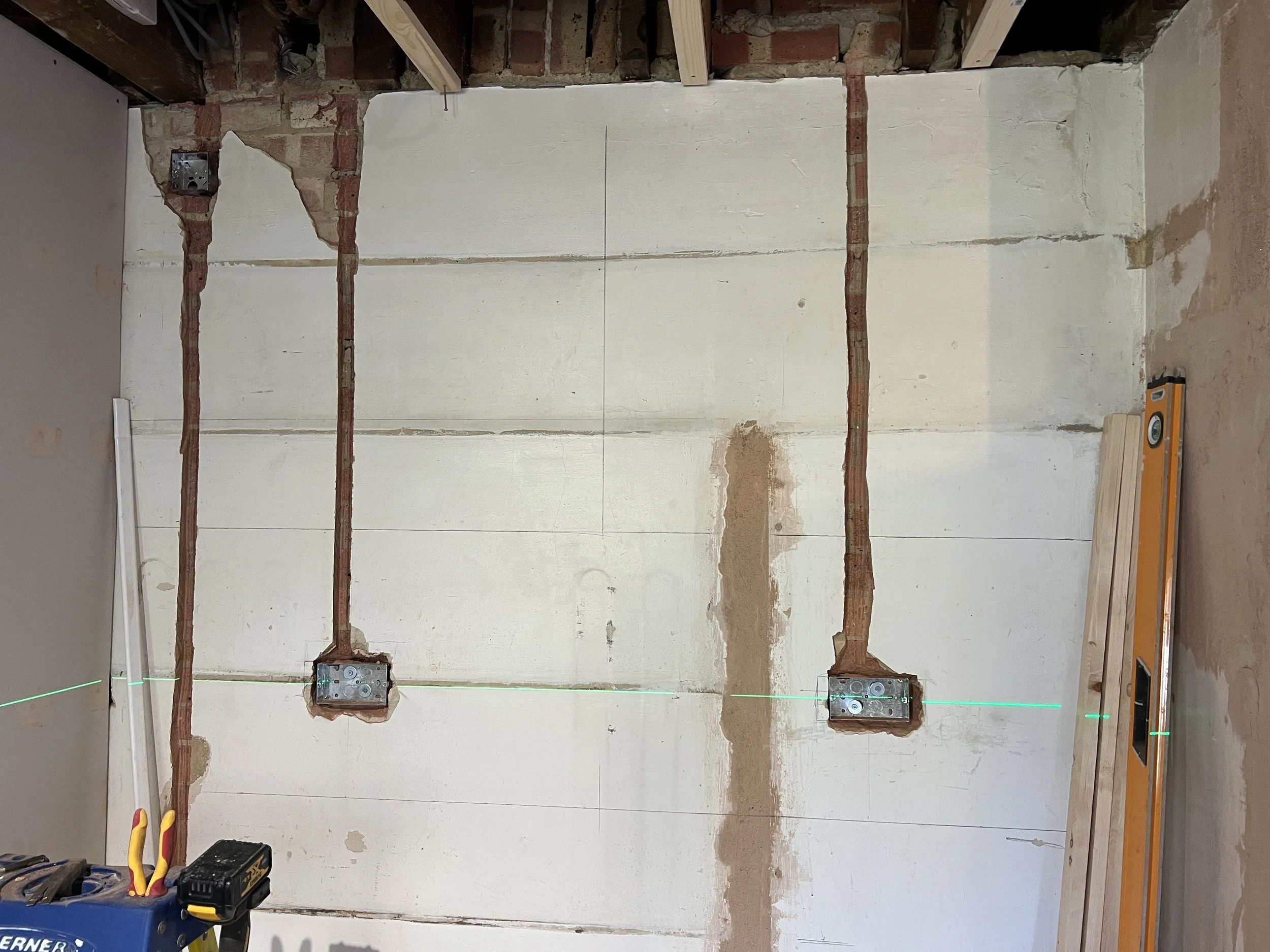 Construction site with electrical wiring in wall, green laser level line, and construction tools on the floor.