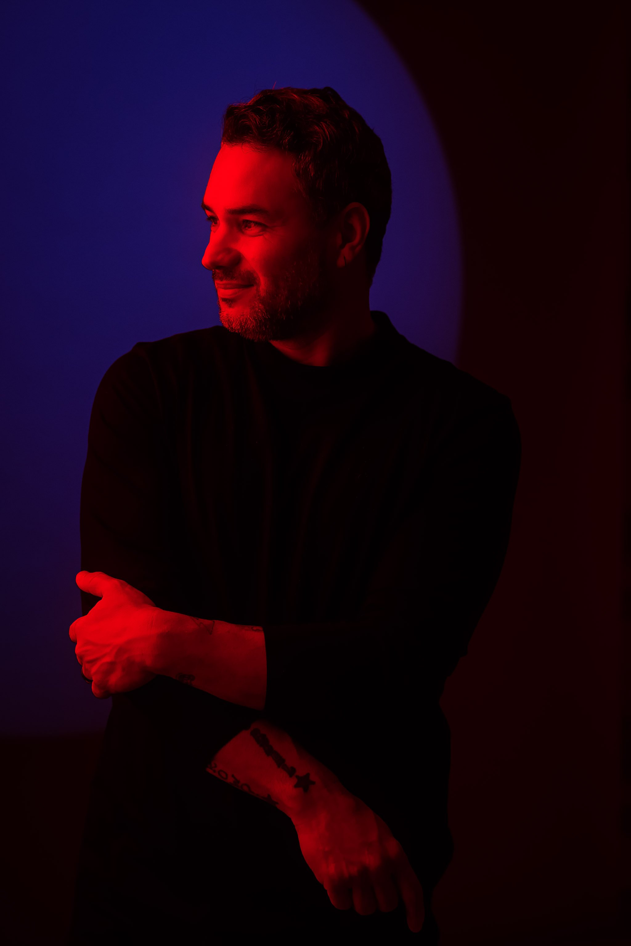 Side profile of person with short dark hair and a beard, smiling with arms crossed, illuminated by red and blue lights, against a dark background.