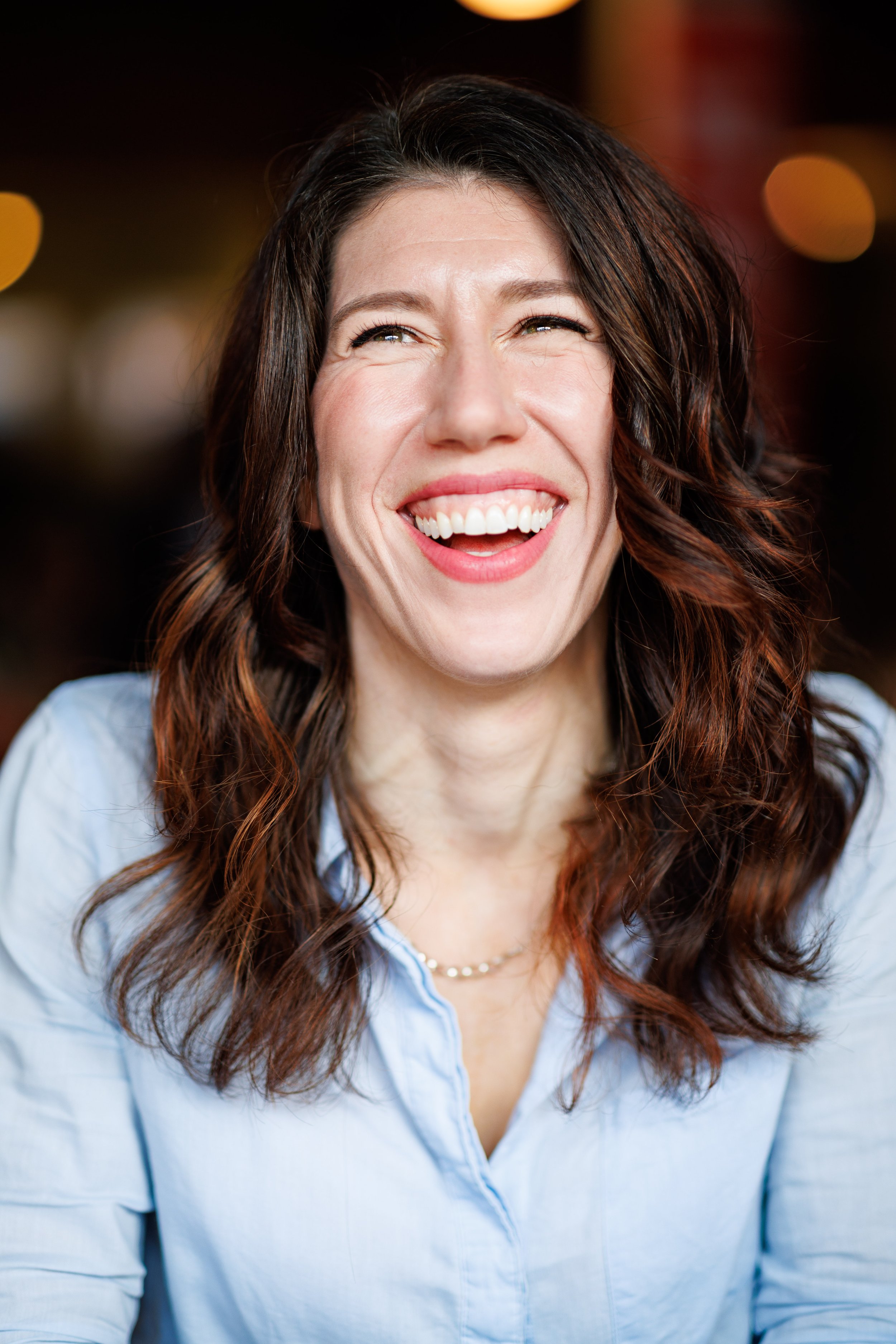 Close-up of a person smiling with visible teeth, wearing a light blue button-up shirt and a necklace, with dark brown wavy hair and soft lighting.