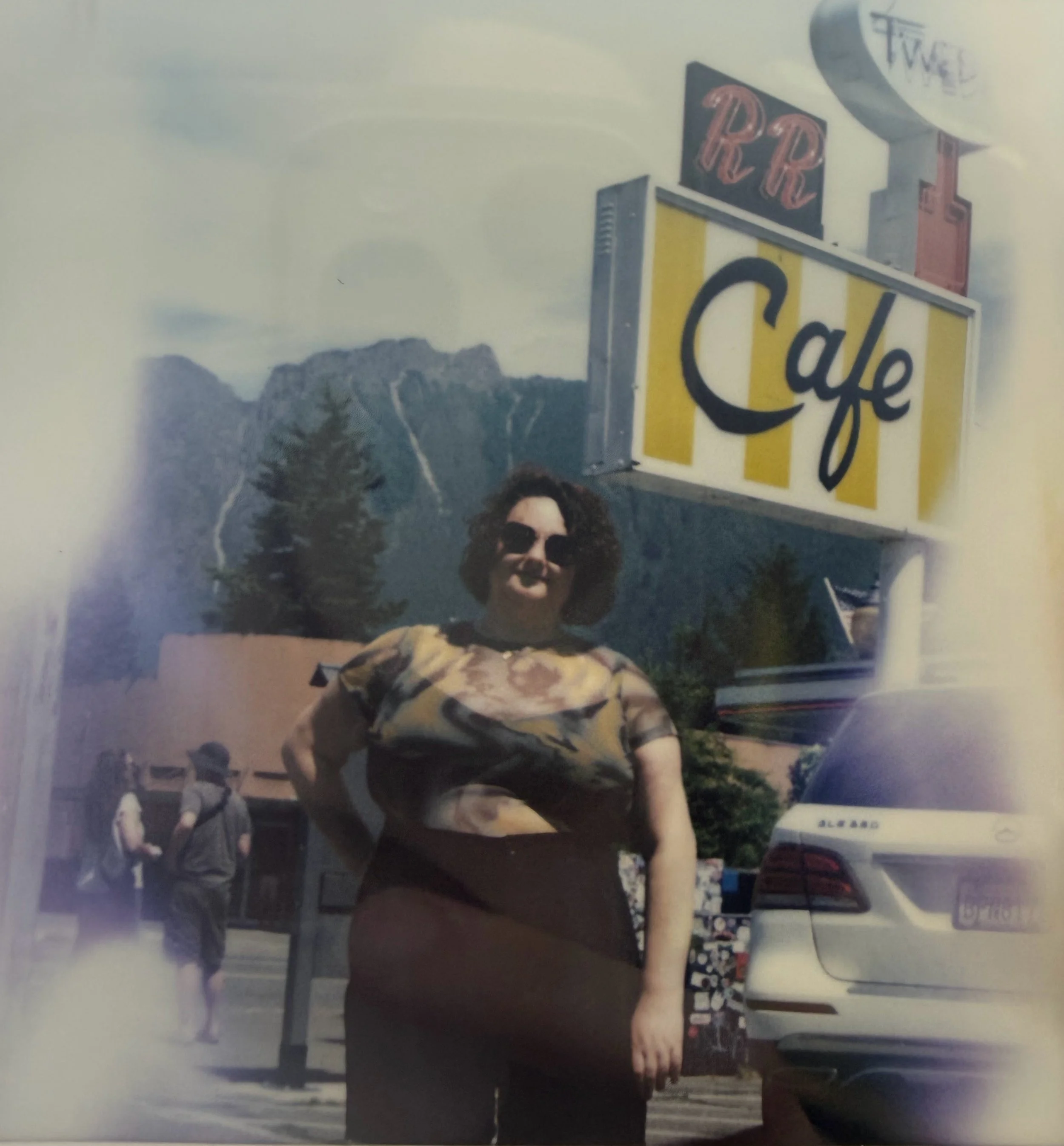 A person with curly hair, wearing glasses and a patterned shirt, standing near a cafe sign with a mountain landscape in the distance.