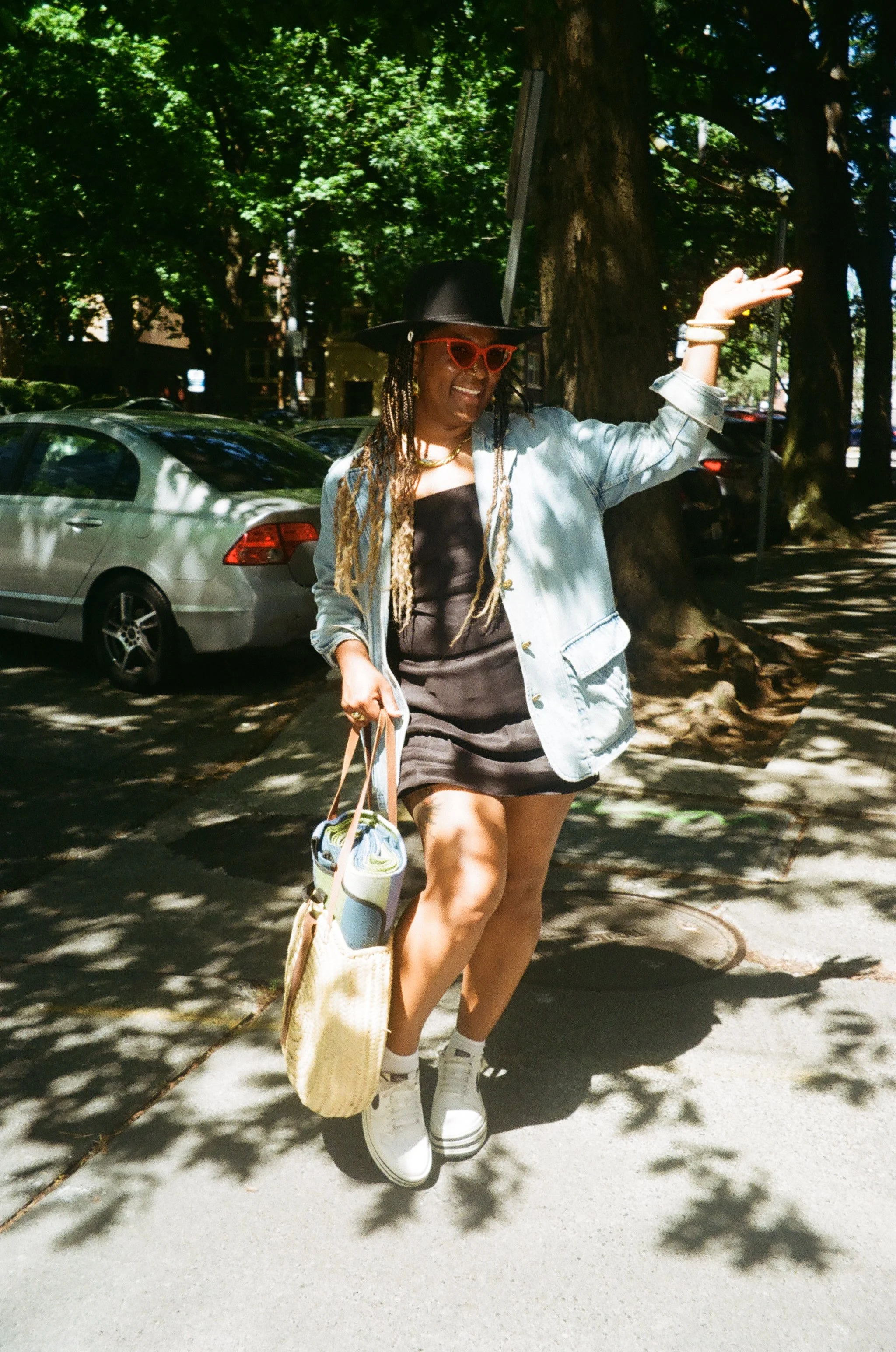 A person with braids wearing a black hat, red sunglasses, a black onesie, and a denim jacket, walking outdoors on a sunny day, carrying a tote bag and a rolled-up blanket.