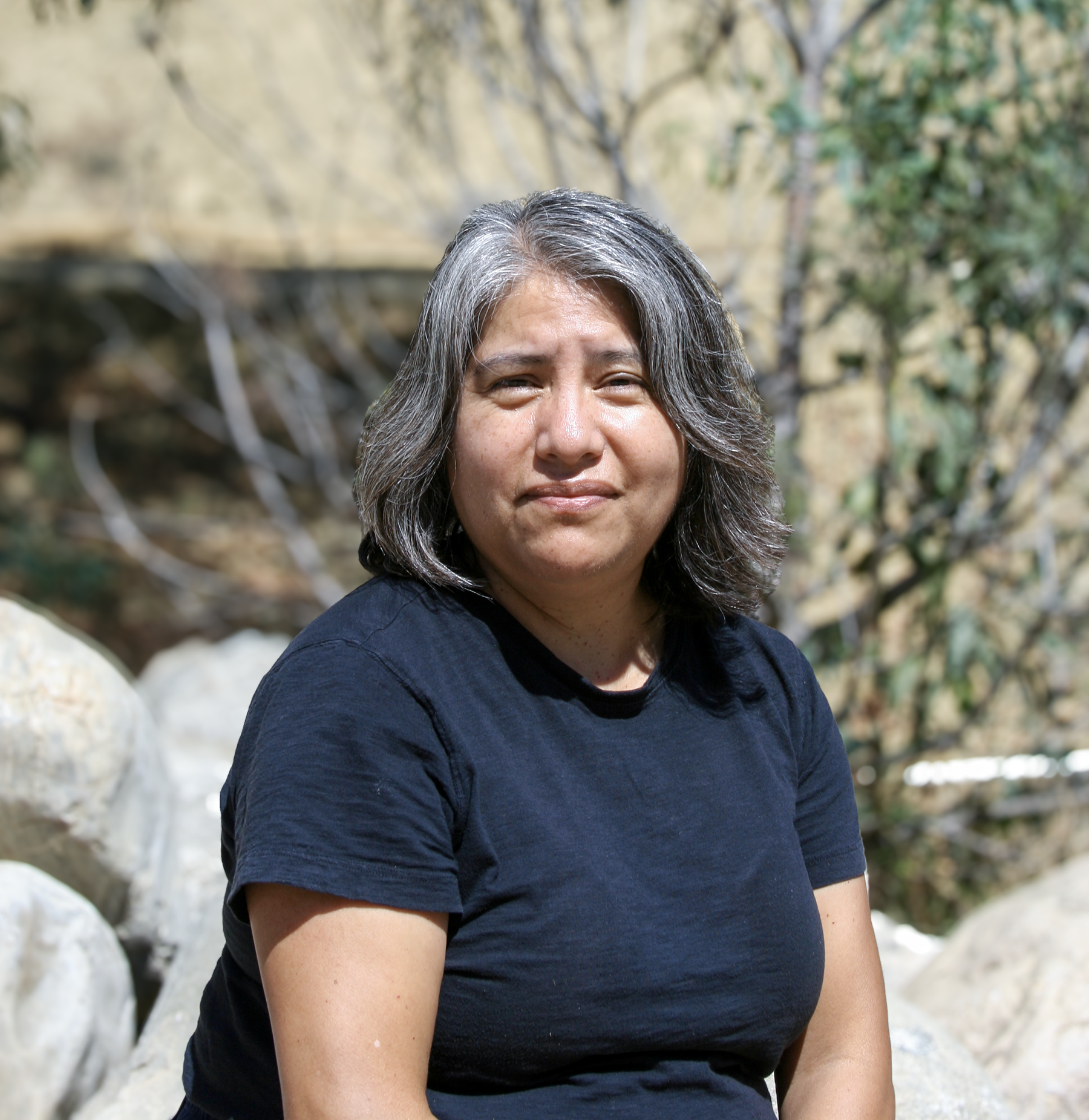 Close-up of a person with shoulder-length salt-and-pepper hair, wearing a black t-shirt, sitting outdoors near rocks and greenery, smiling gently.