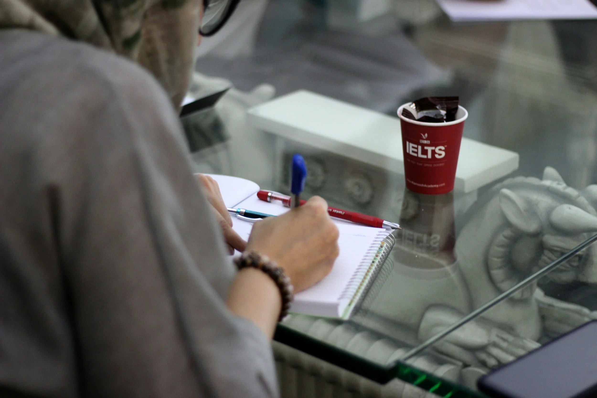 A person writing in a notebook at a glass desk with a red IELTS cup and chocolate bars on the desk.