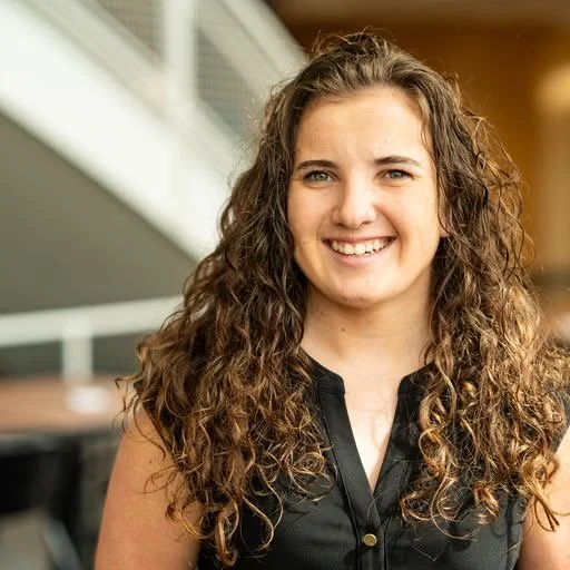 A young woman with long, curly brown hair and blue eyes, smiling, in an indoor setting.
