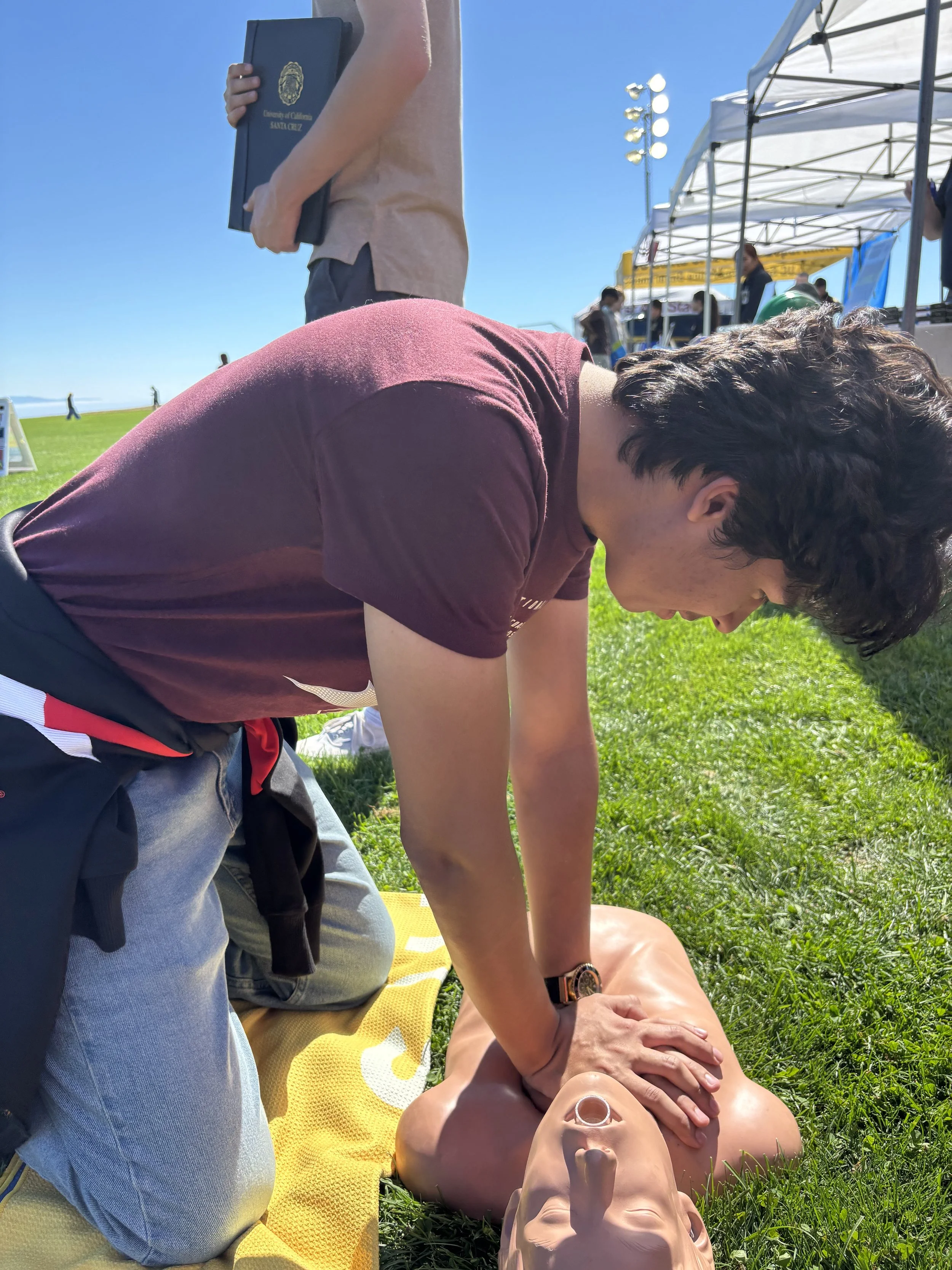 Person performing CPR on a mannequin outdoors during daytime training session.