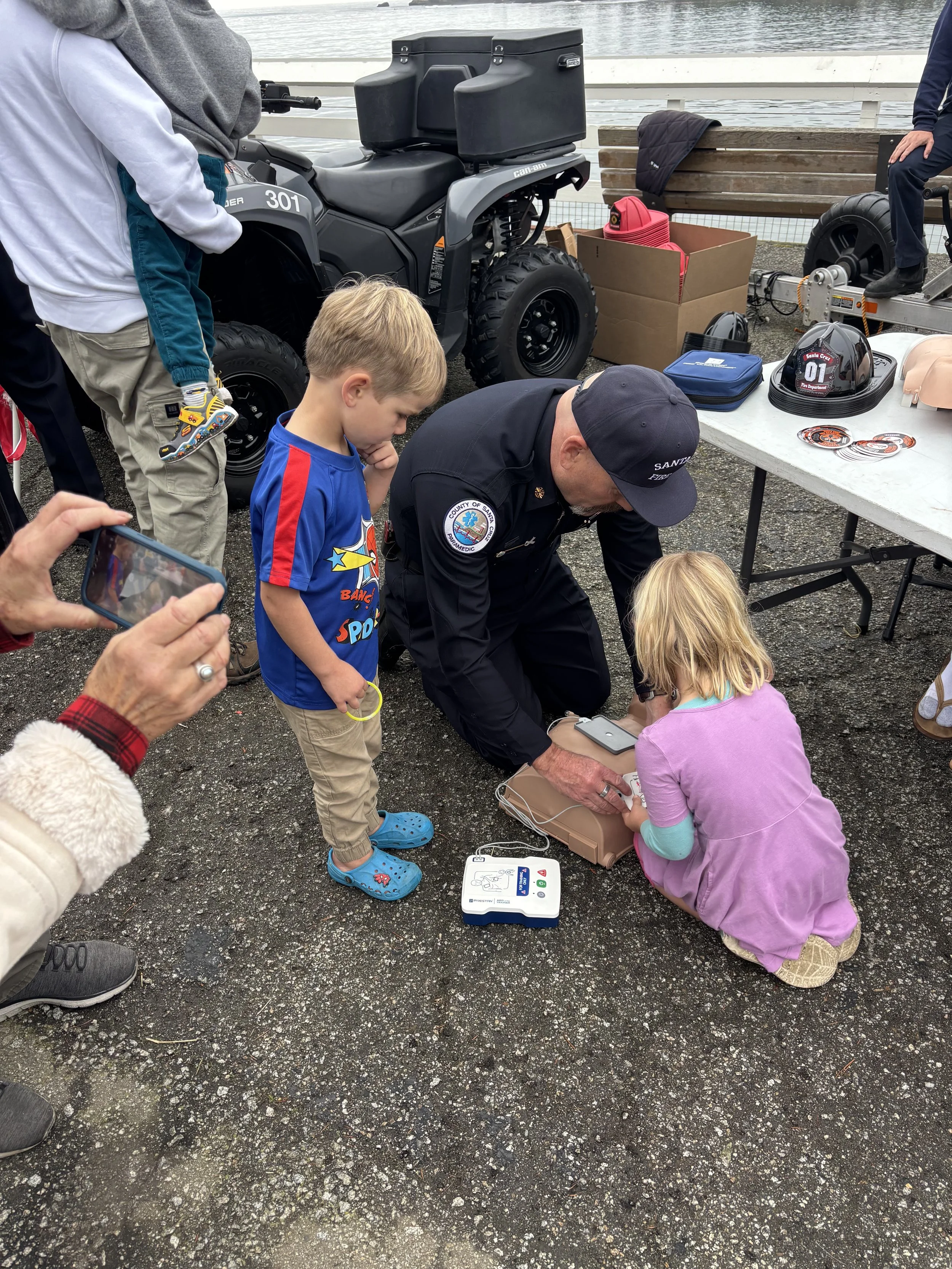 A firefighter demonstrates an emergency medical device to children at an outdoor event by the water, with equipment and a small ATV in the background.