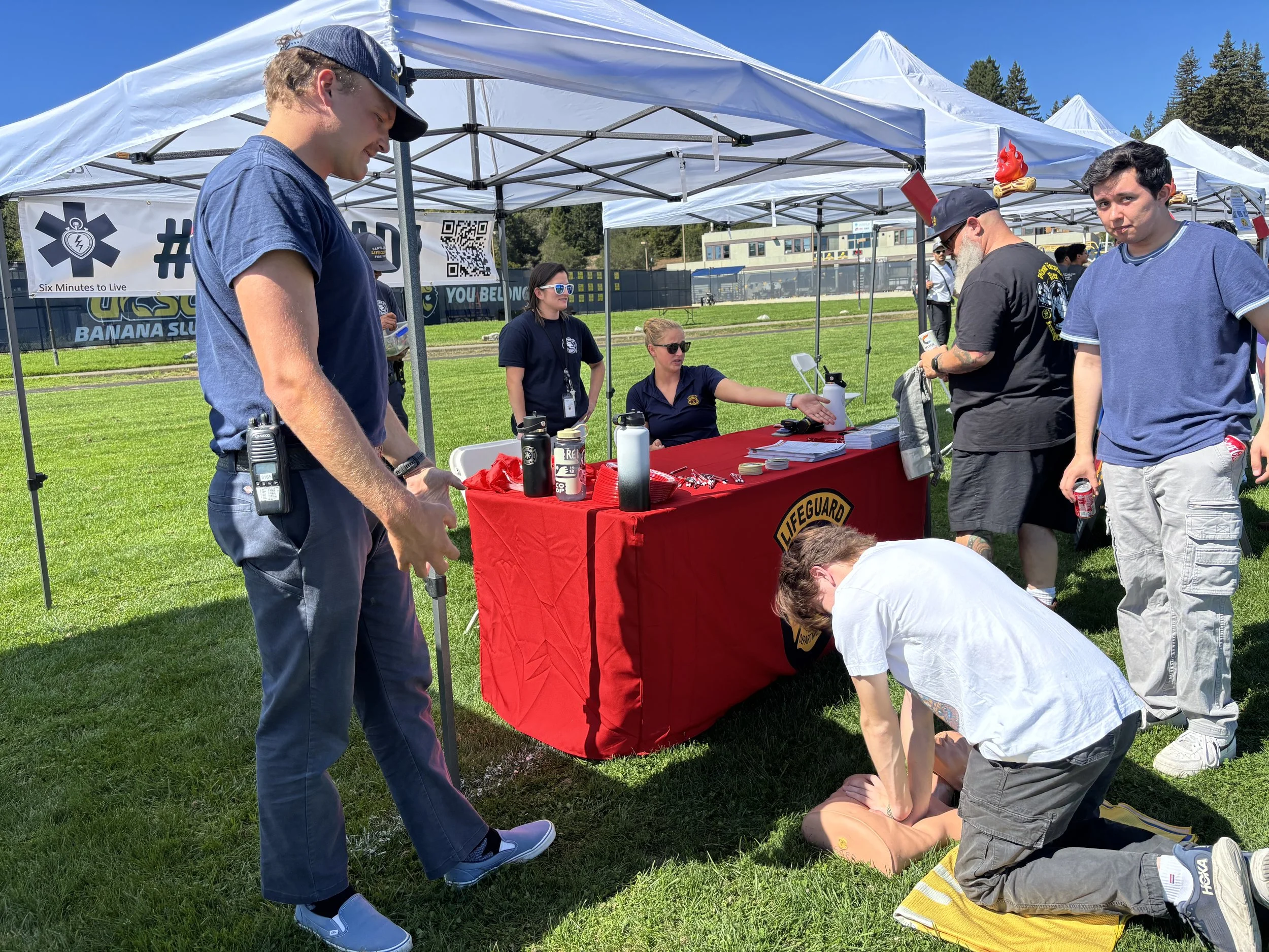 Lifeguards and emergency responders perform CPR on a mannequin at an outdoor event, with tents, banners, and people observing.