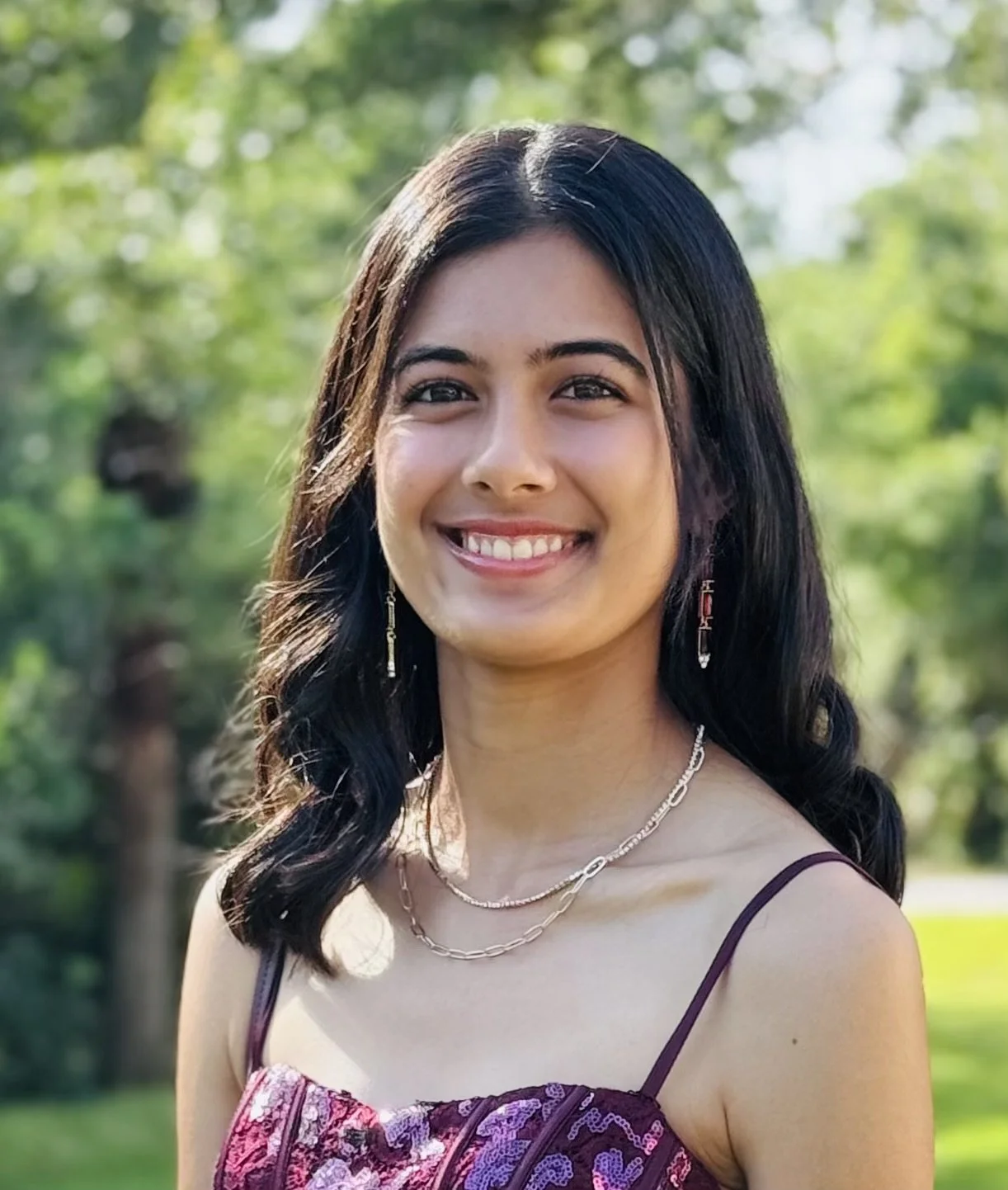 A smiling woman with dark hair, wearing a purple dress, earrings, and layered necklaces, standing outdoors in a green park with trees in the background.