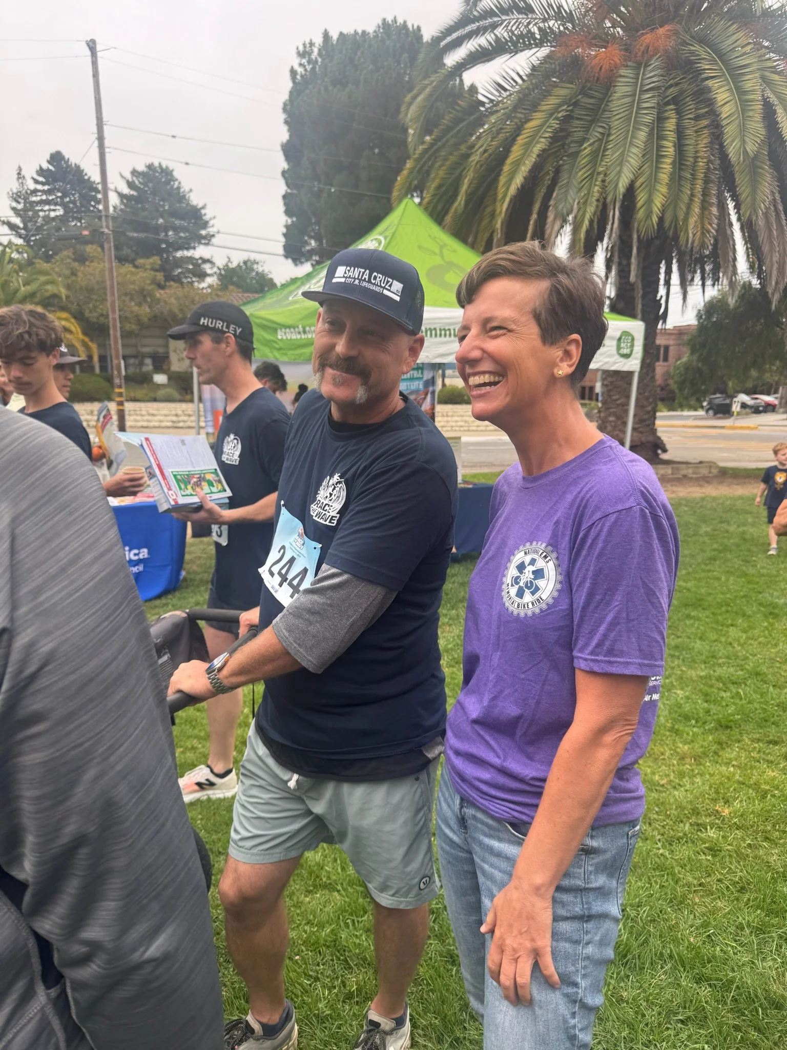 Two smiling people, a man and a woman, standing outdoors at a community event or race. The man is wearing a black cap, navy T-shirt, gray shorts, and is pushing a stroller. The woman has short hair, is wearing a purple T-shirt with a logo, and jeans. There are other people and a green tent in the background, with trees and a palm tree visible.