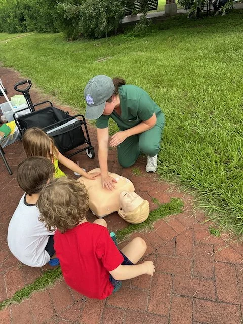 A woman demonstrating CPR on a mannequin while three children watch outdoors on a brick sidewalk.