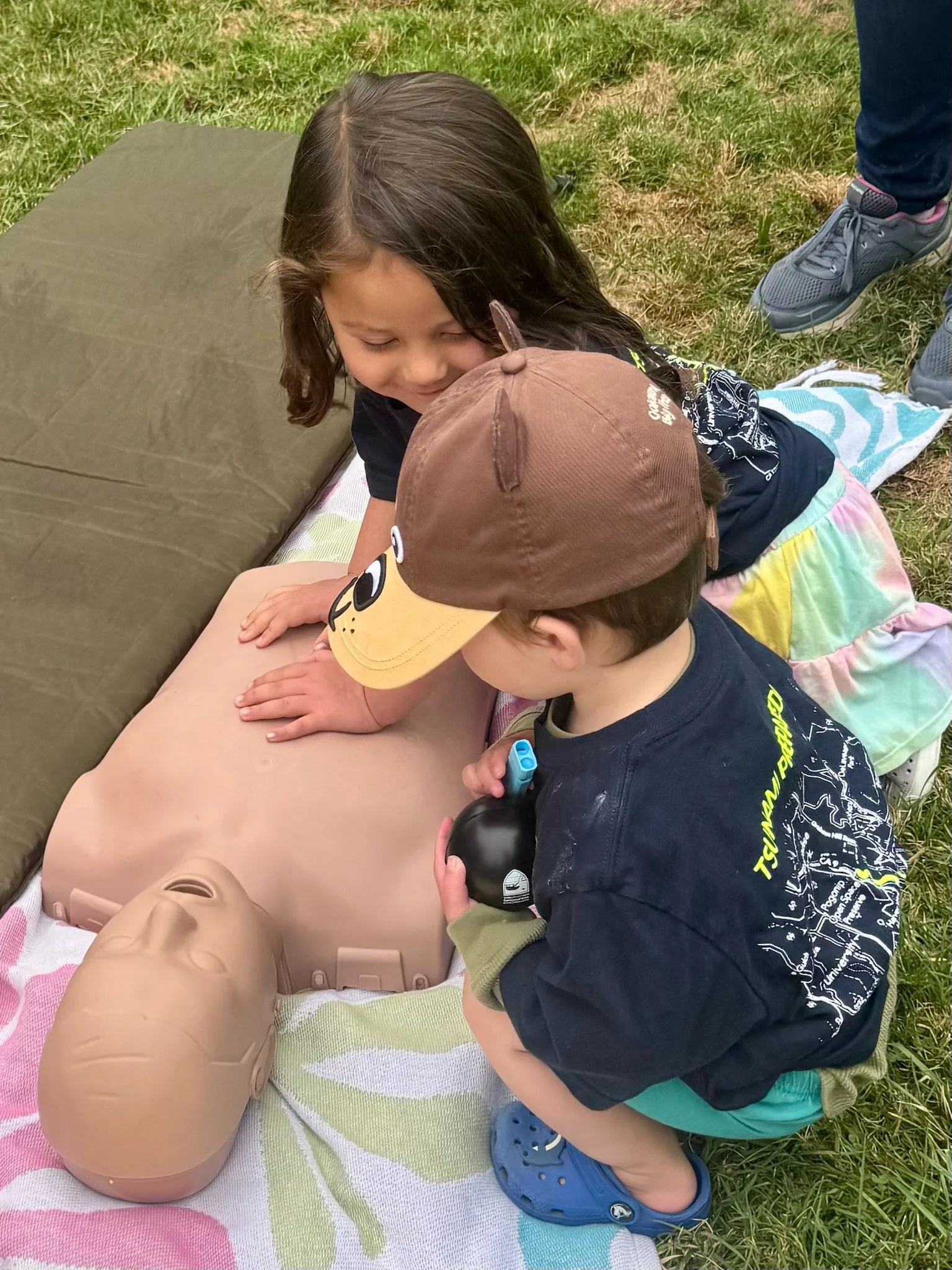Two children practicing CPR on a training dummy outdoors, with one child performing chest compressions and the other observing.