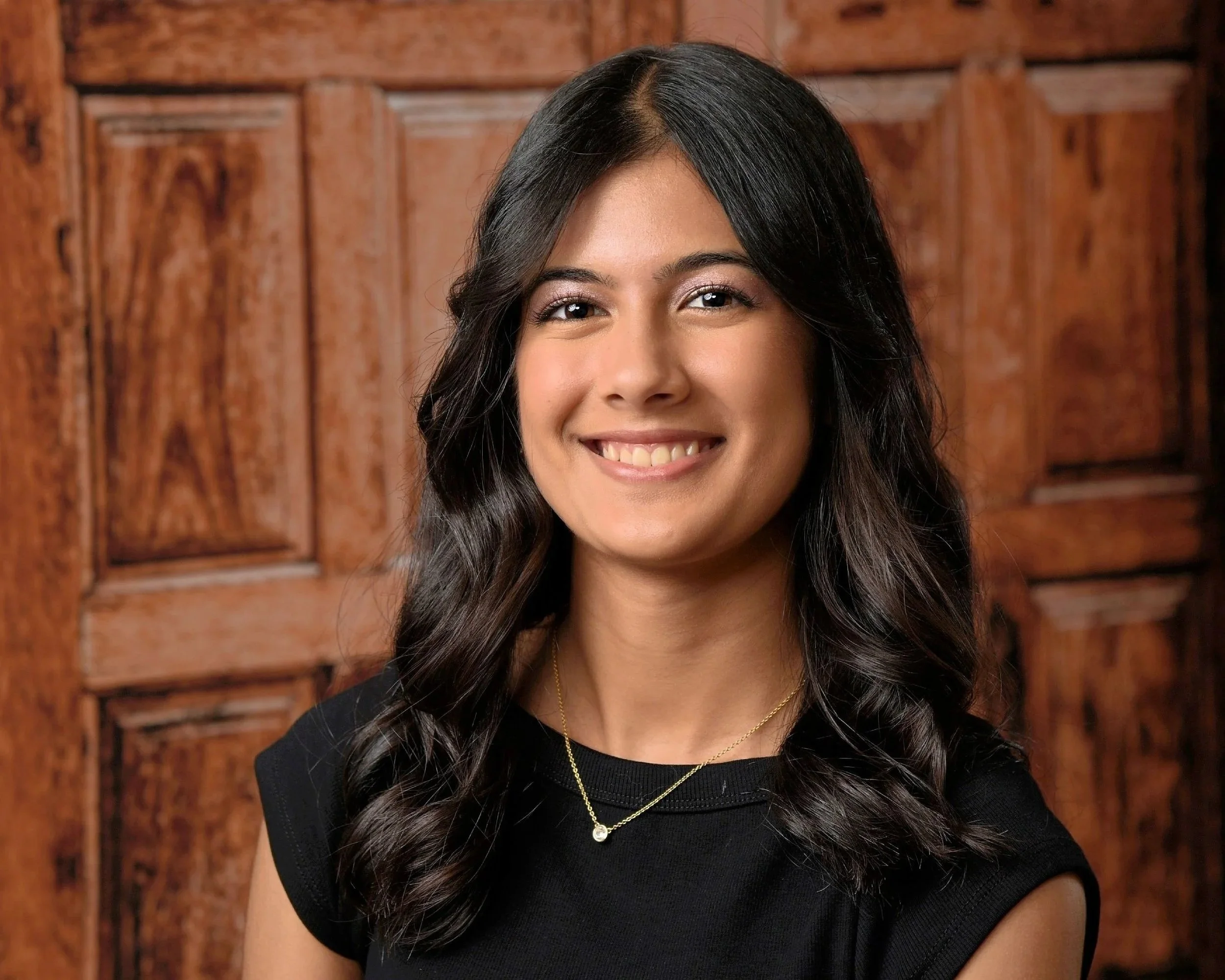 A smiling young woman with long wavy dark hair, wearing a black top and a gold necklace with a small pendant, standing in front of a wooden door or paneling.
